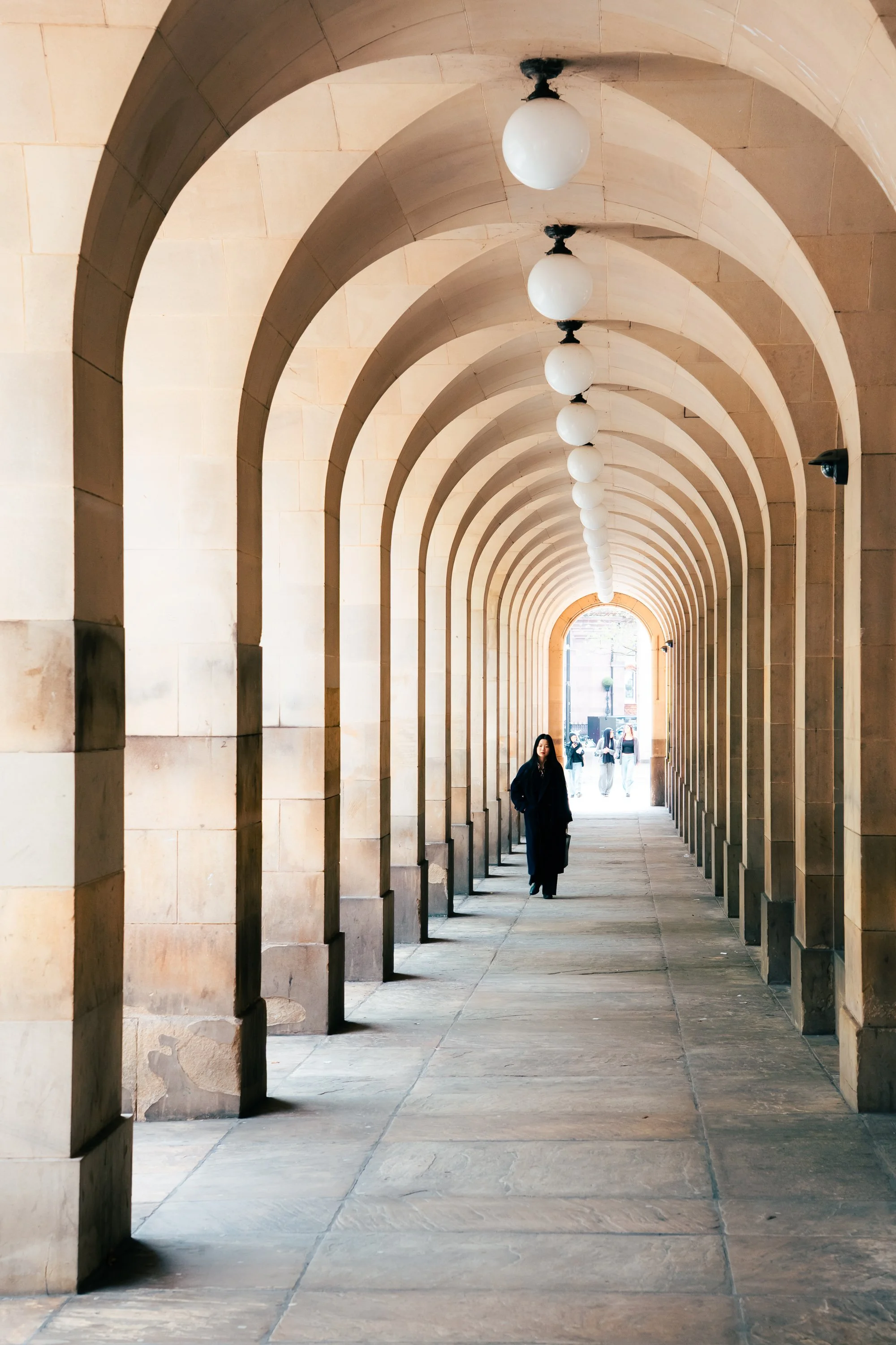 A stone walking corridor with arches and hanging spherical lights, with a person walking towards the camera and other people in the background.