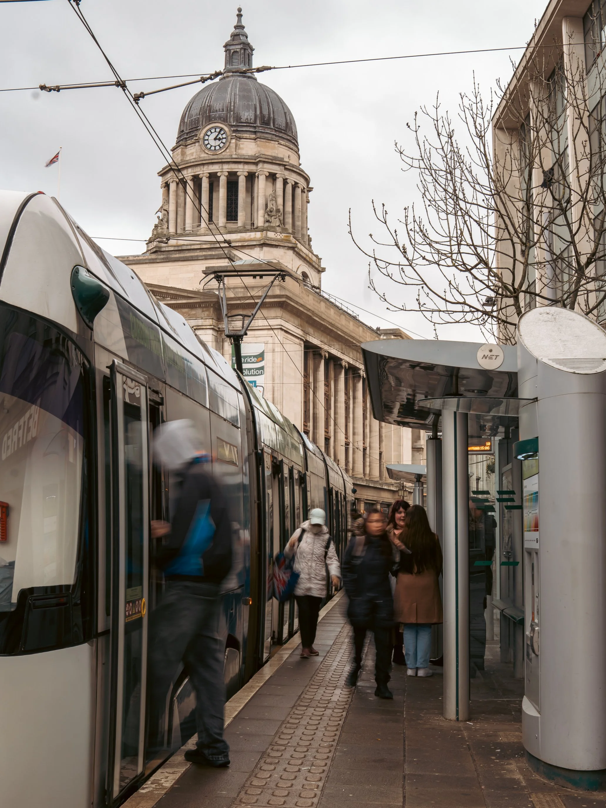 People waiting at a tram or bus stop with a modern shelter and a historic building with a clock tower in the background.