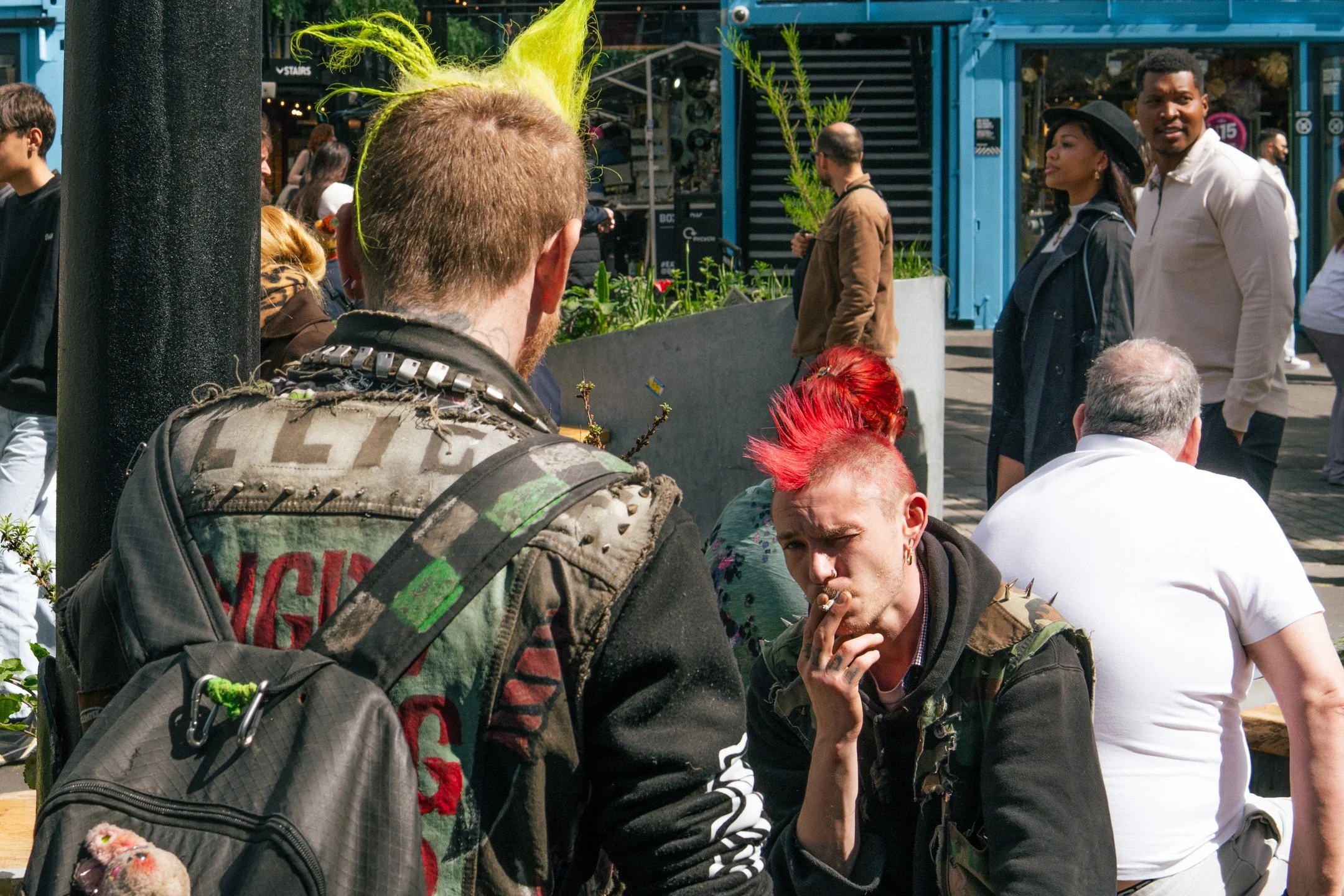 A crowd of people gathered on a city street with some individuals with brightly colored hairstyles and clothing, including a person with bright green hair and others with red hair and punk-inspired fashion.