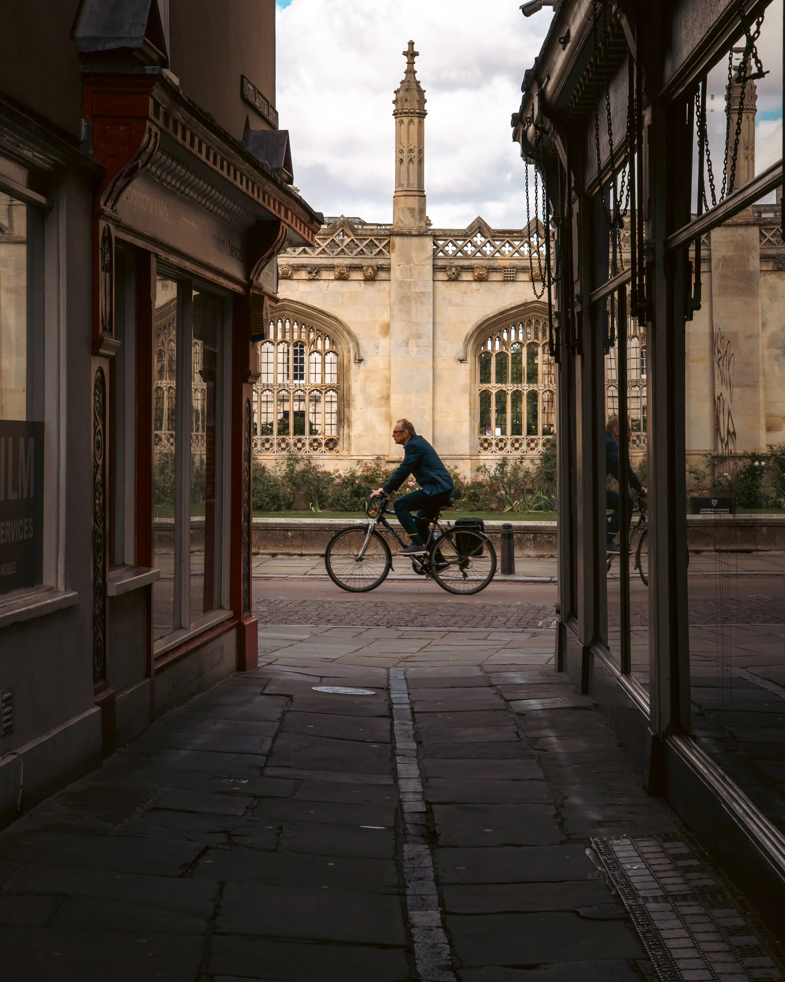 A cyclist riding a bicycle through a narrow passageway between black storefronts with a historic building in the background, featuring a large gothic-style window and a tall decorative tower, under a cloudy sky.