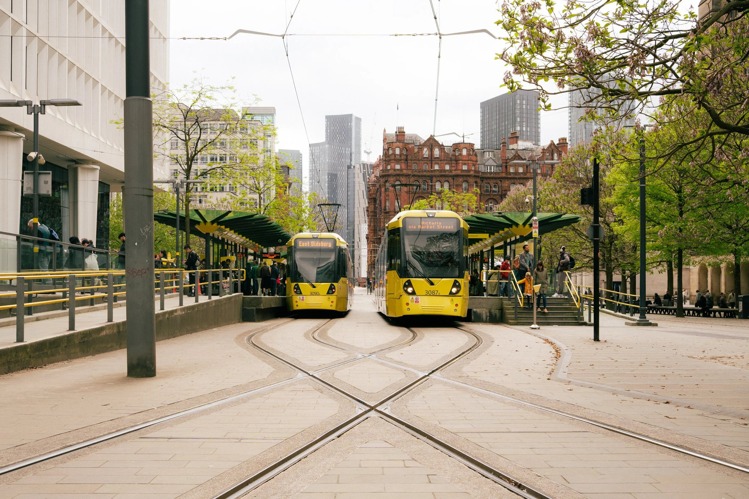 Two yellow trams at a city tram station with buildings, trees, and people waiting or walking nearby.