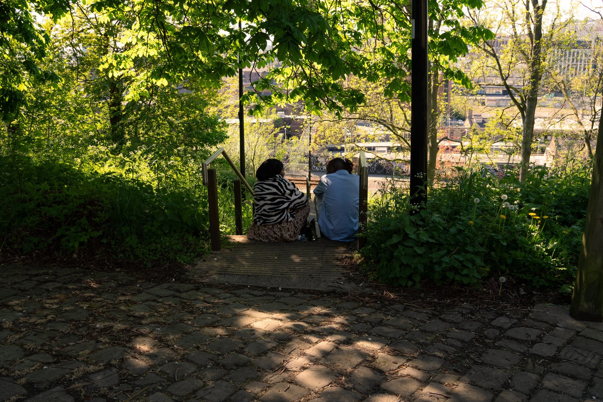 Two people sitting on a bench in a shaded park surrounded by green trees, with urban buildings visible in the background.
