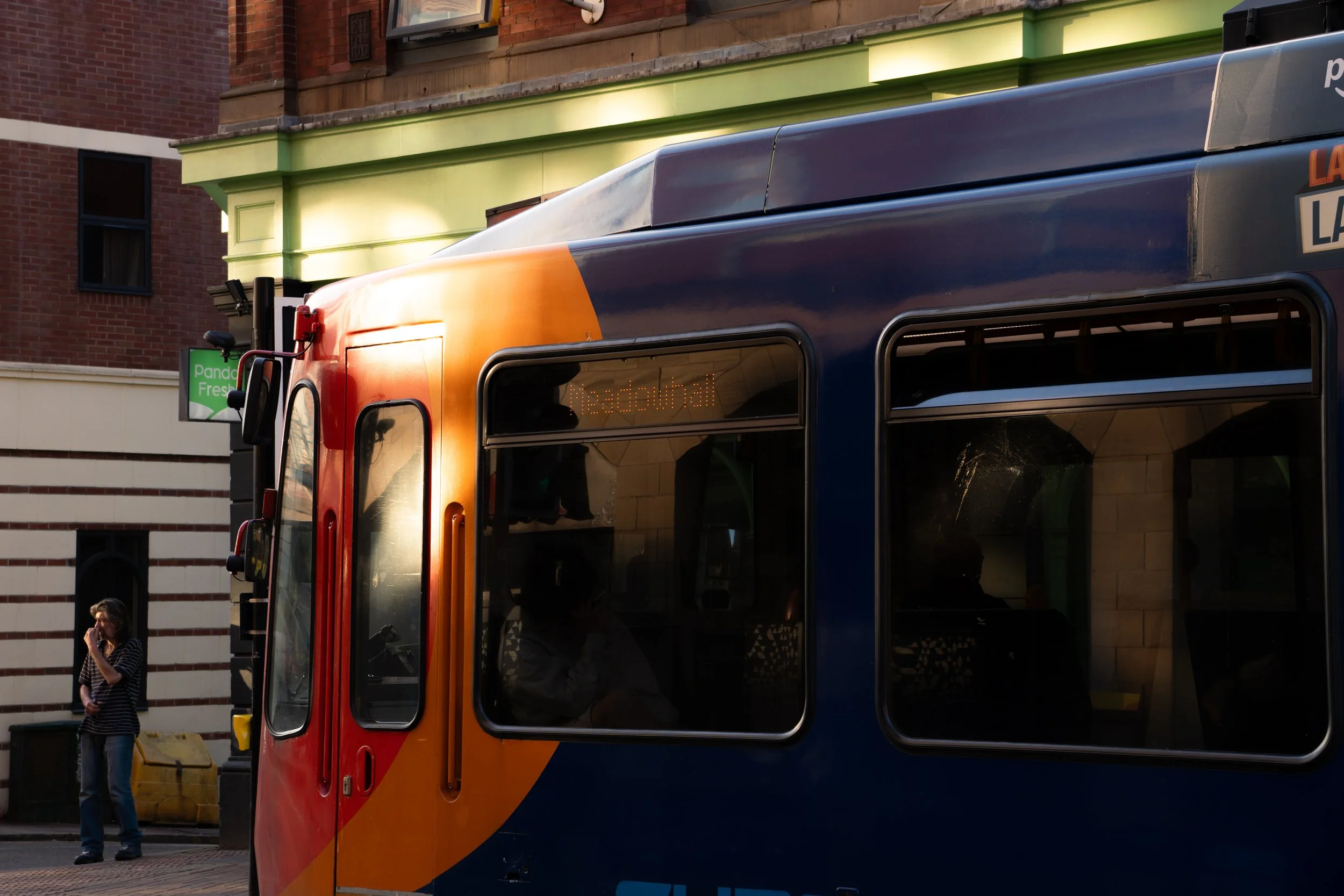 A colorful tram with orange and blue colors traveling on a city street, with a woman talking on her phone and a man walking nearby. The background includes brick buildings and a green storefront.