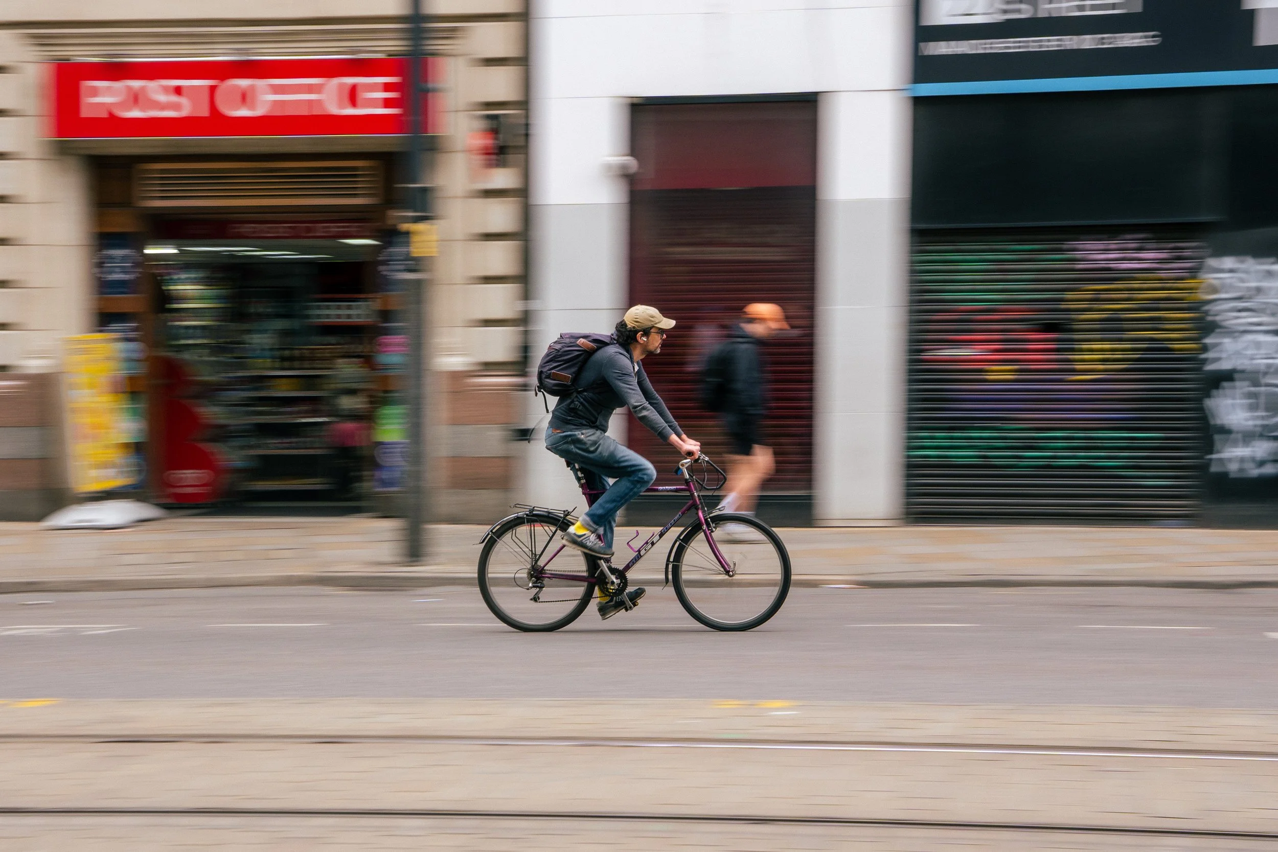 A man riding a bicycle on a city street with storefronts and advertisements in the background.