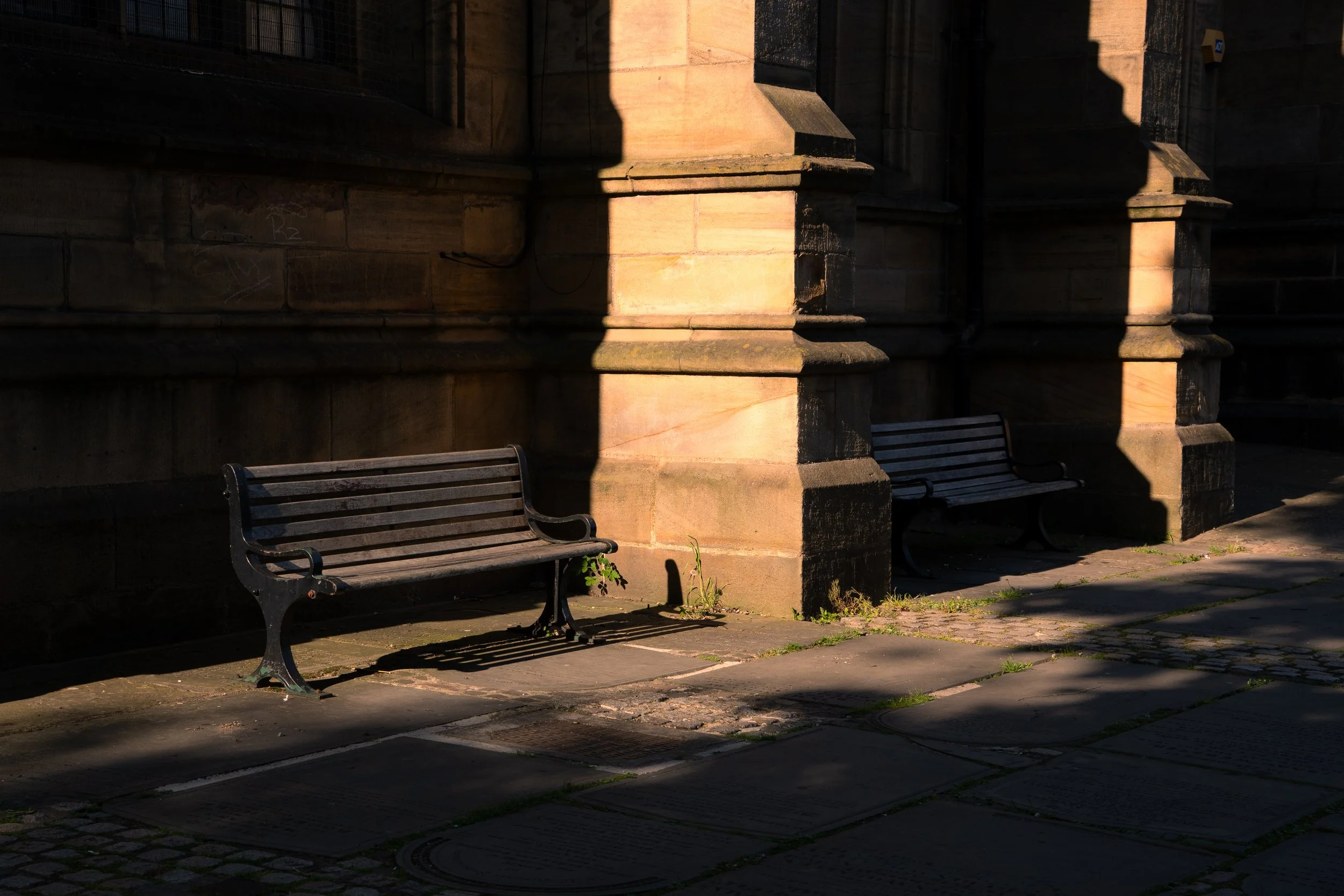Sunlit stone building with two empty park benches beneath shaded areas on a sidewalk.
