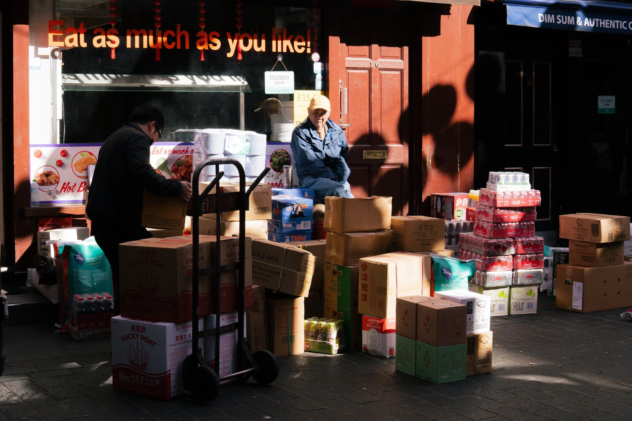 Street vendor's shop with stacked boxes of food and drinks, a sign reading 'Eat as much as you like!', and two people sitting outside under sunlight.