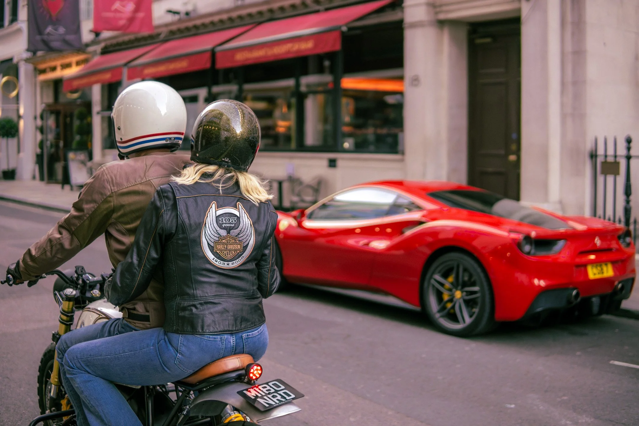 Two people riding a motorcycle on a city street. They wear helmets, and one has a Harley-Davidson jacket. In the background, there is a red sports car and storefronts with red awnings.