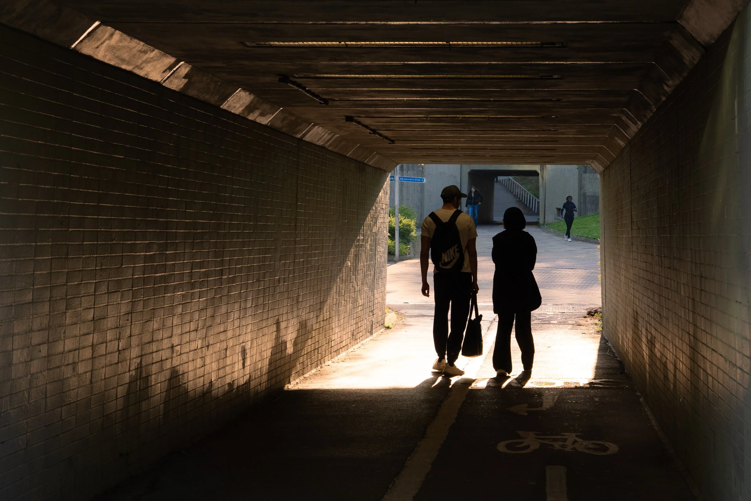People walking through a tunnel, with sunlight illuminating the exit areas. A bicycle lane is visible on the ground inside the tunnel.