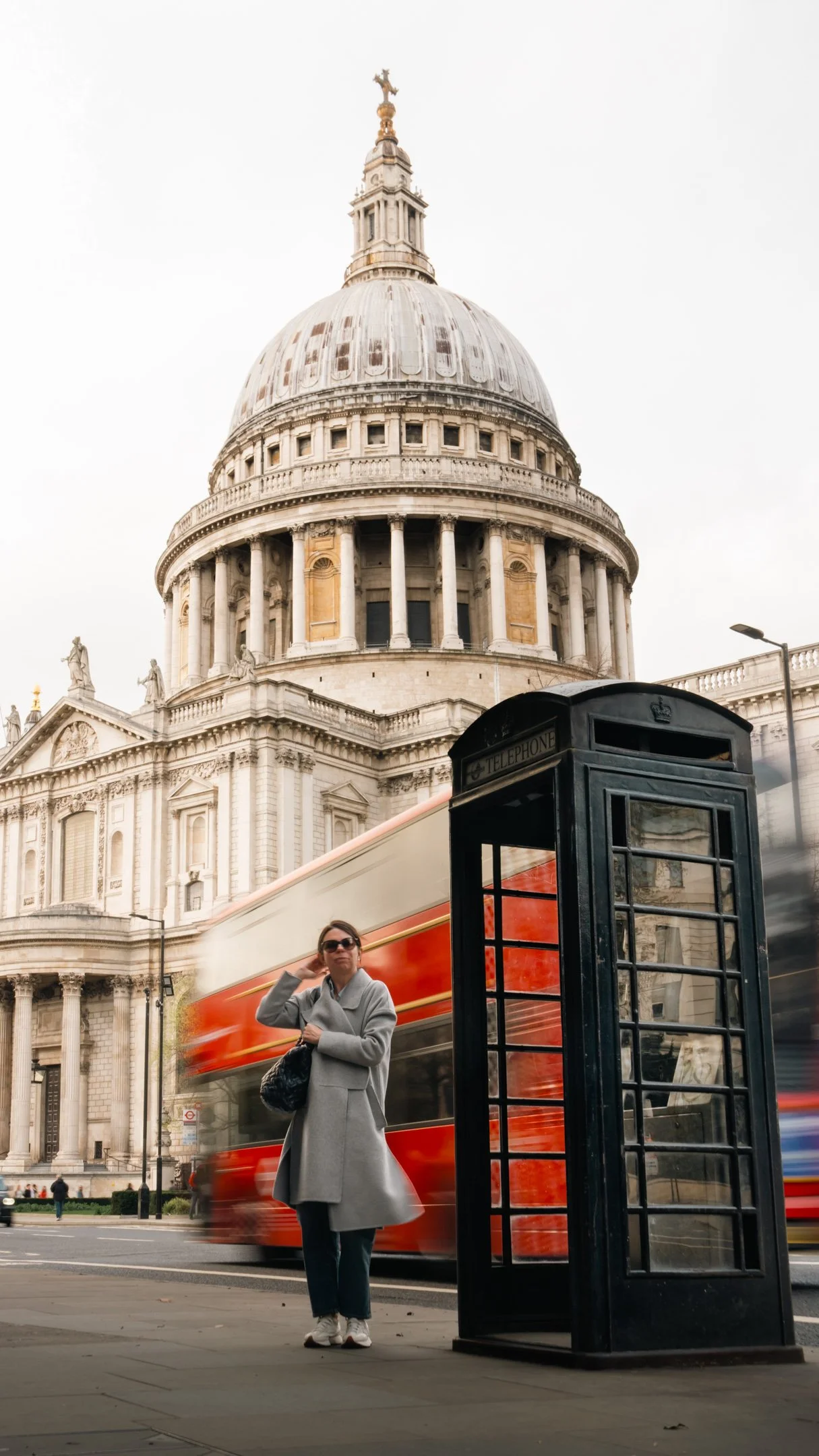A woman in a gray coat and sunglasses standing near a black telephone booth on a city street, with a red double-decker bus passing by and St. Paul's Cathedral in London in the background.