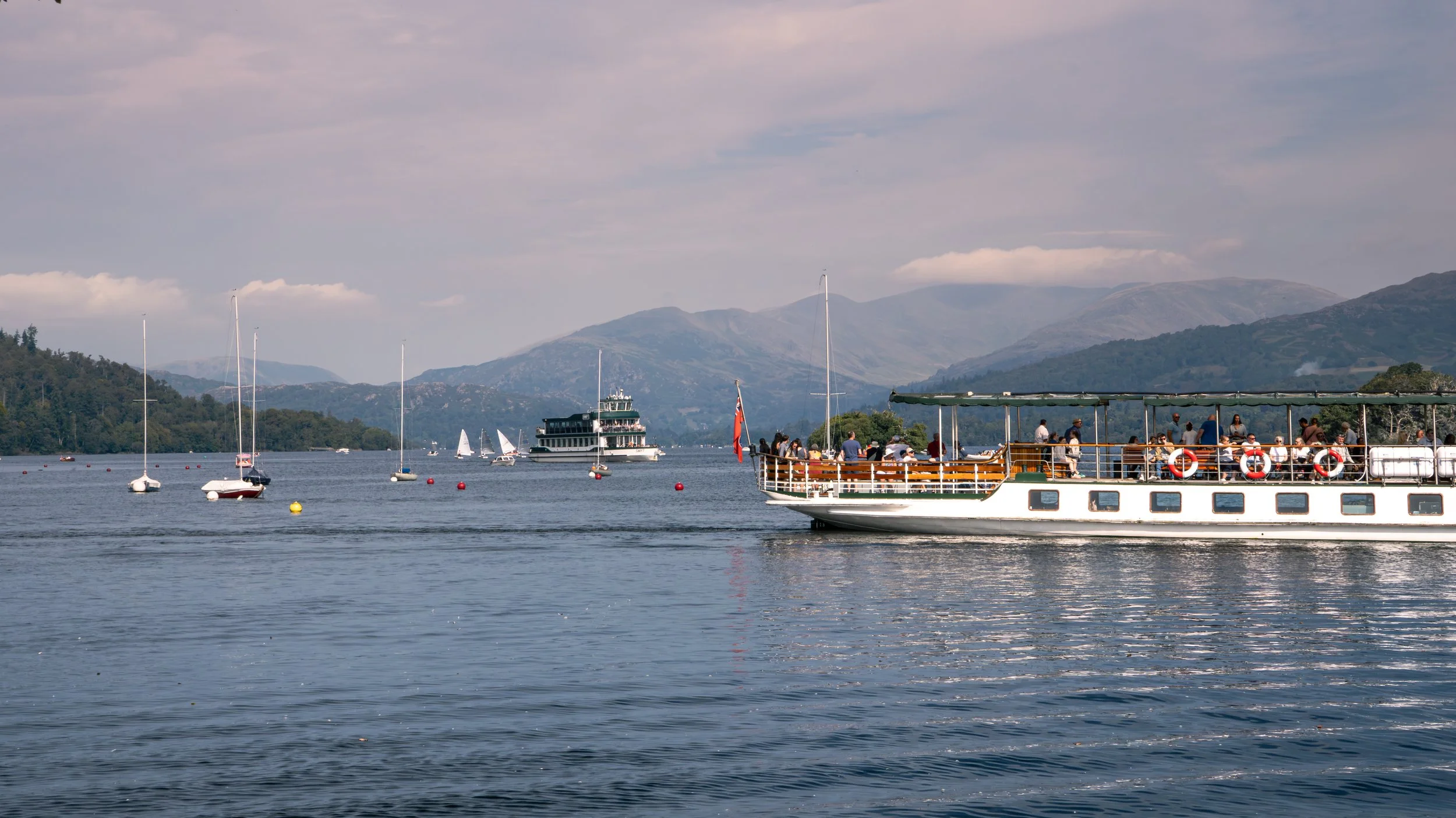 People enjoying a boat cruise on a lake with sailboats and a large sightseeing boat, surrounded by green hills and mountains under a partly cloudy sky.