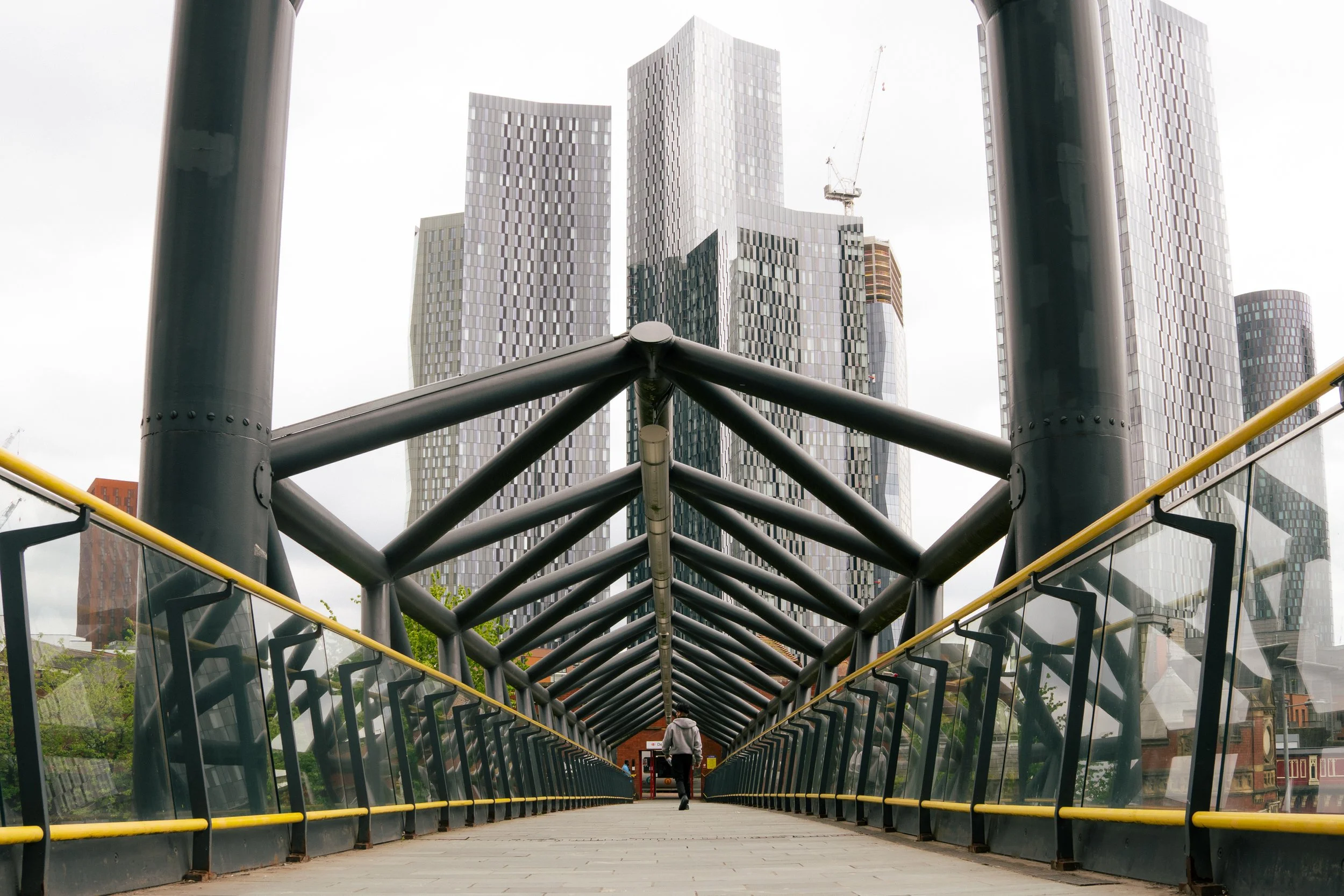 A person walking on a glass-covered pedestrian bridge with metal framework, with modern skyscrapers in the background.