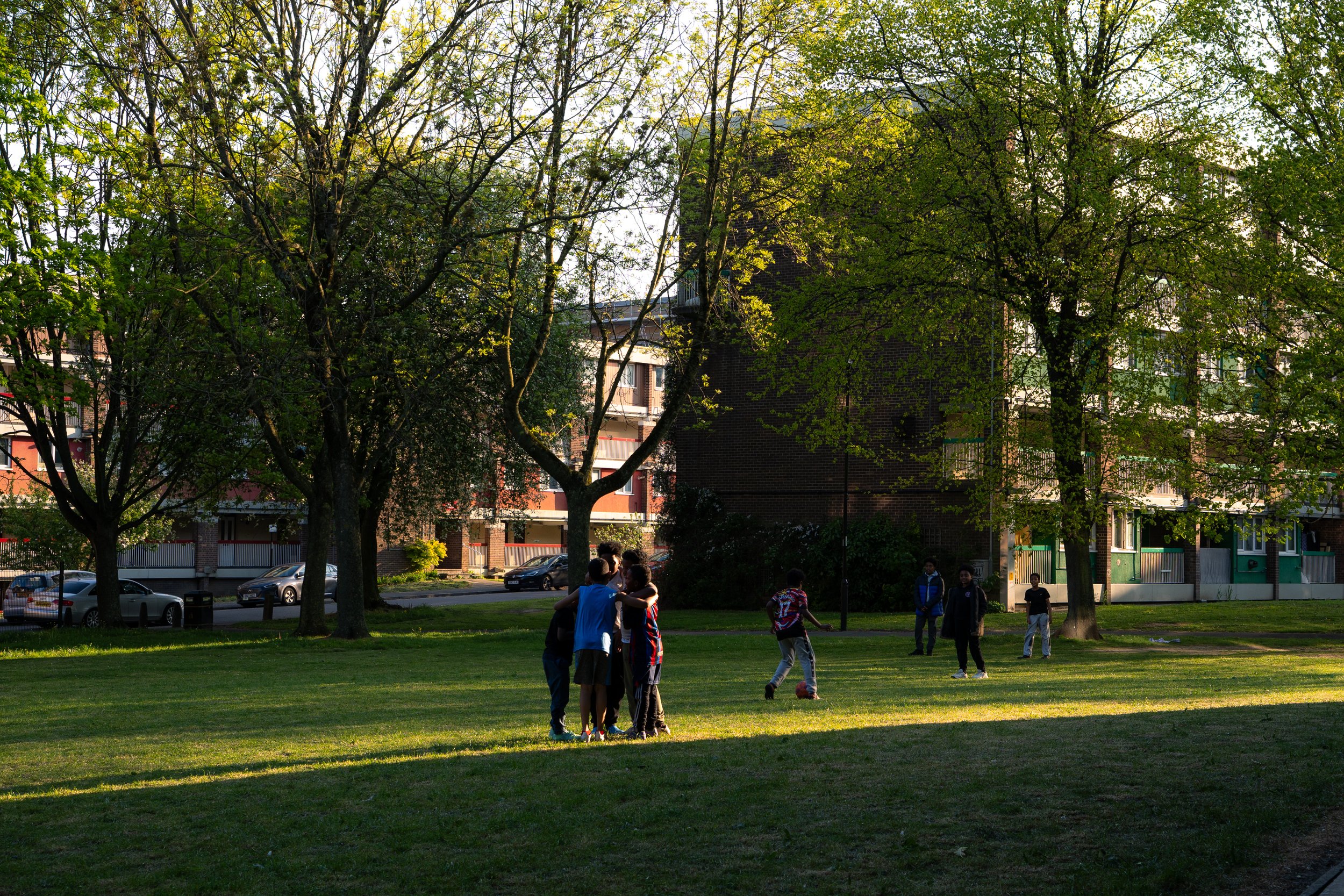 Children playing soccer in a park with trees and apartment buildings in the background during late afternoon or early evening.