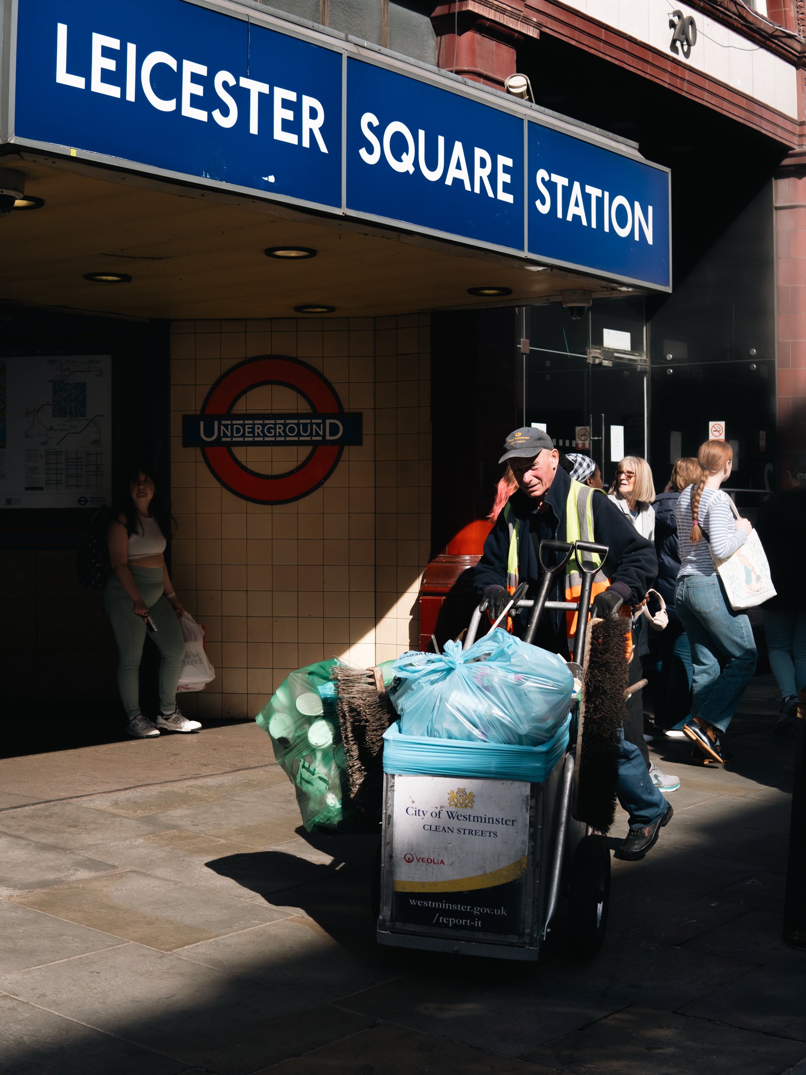 People gathered outside Westminster Underground station, including a man in a high visibility vest with a cart piled with trash bags, and others standing or walking nearby.