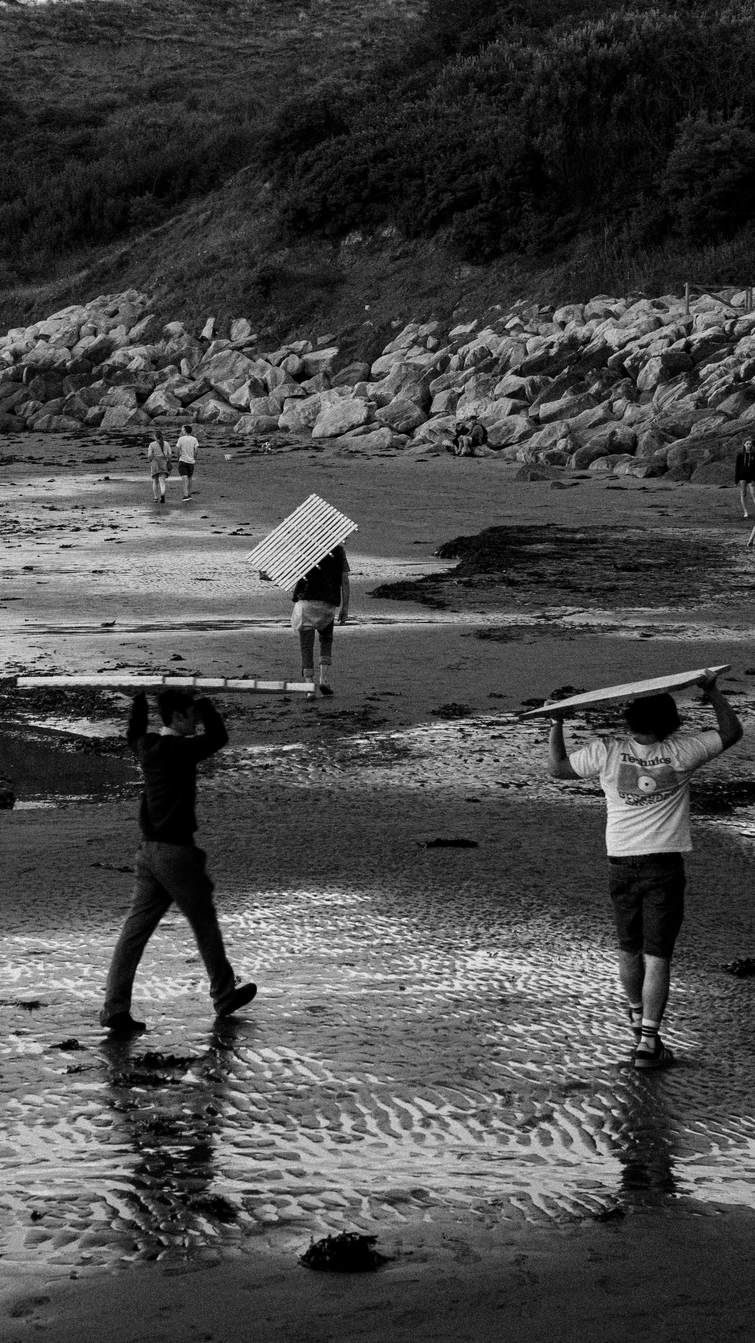 People walking on a beach with some carrying umbrellas, rocks along the shoreline, and a hillside with dense vegetation in the background.