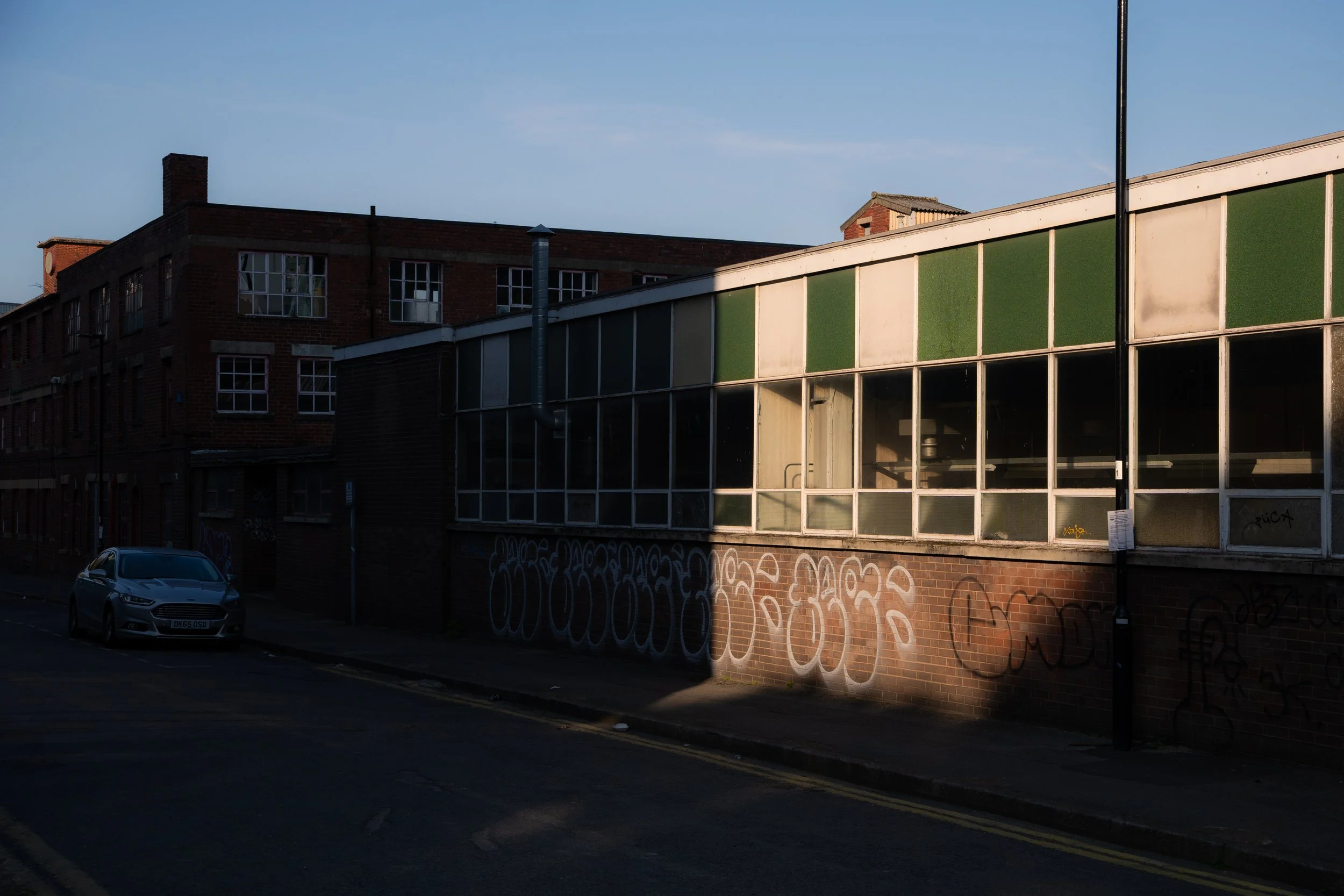 A city street scene at dusk with a white car parked on the side, a brick building with multiple windows, and a graffiti-covered wall under a clear sky.