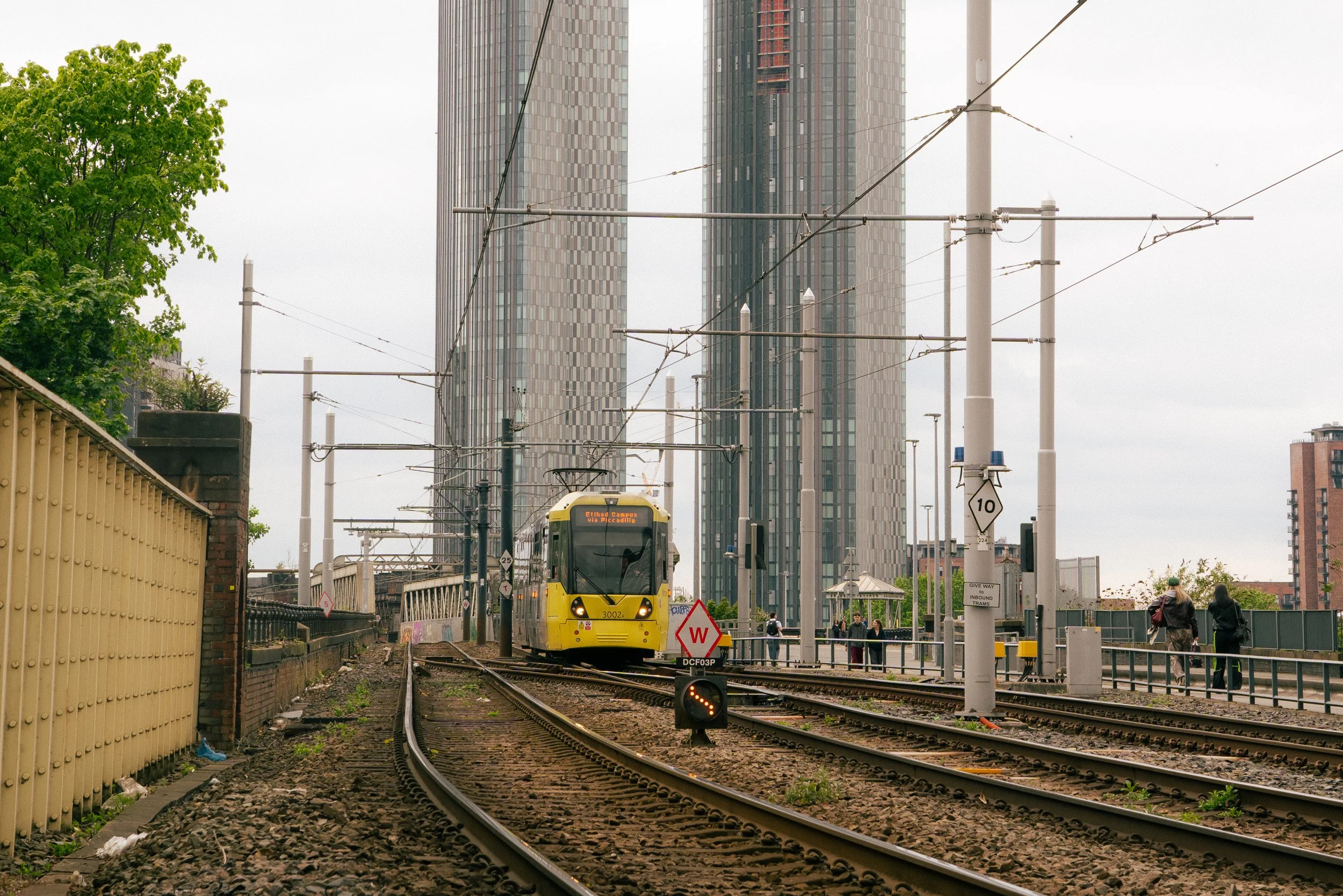 A yellow train traveling down train tracks in an urban area with high-rise buildings in the background and overhead tram wires above.