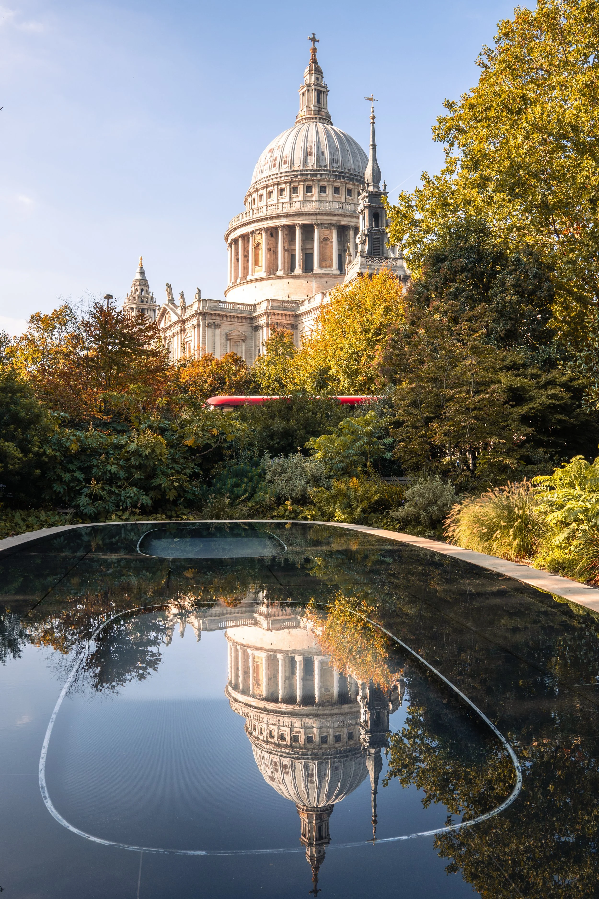 A view of St. Paul's Cathedral in London, with clear blue sky, surrounded by trees with autumn foliage, and a reflective pool of water in the foreground mirroring the cathedral.
