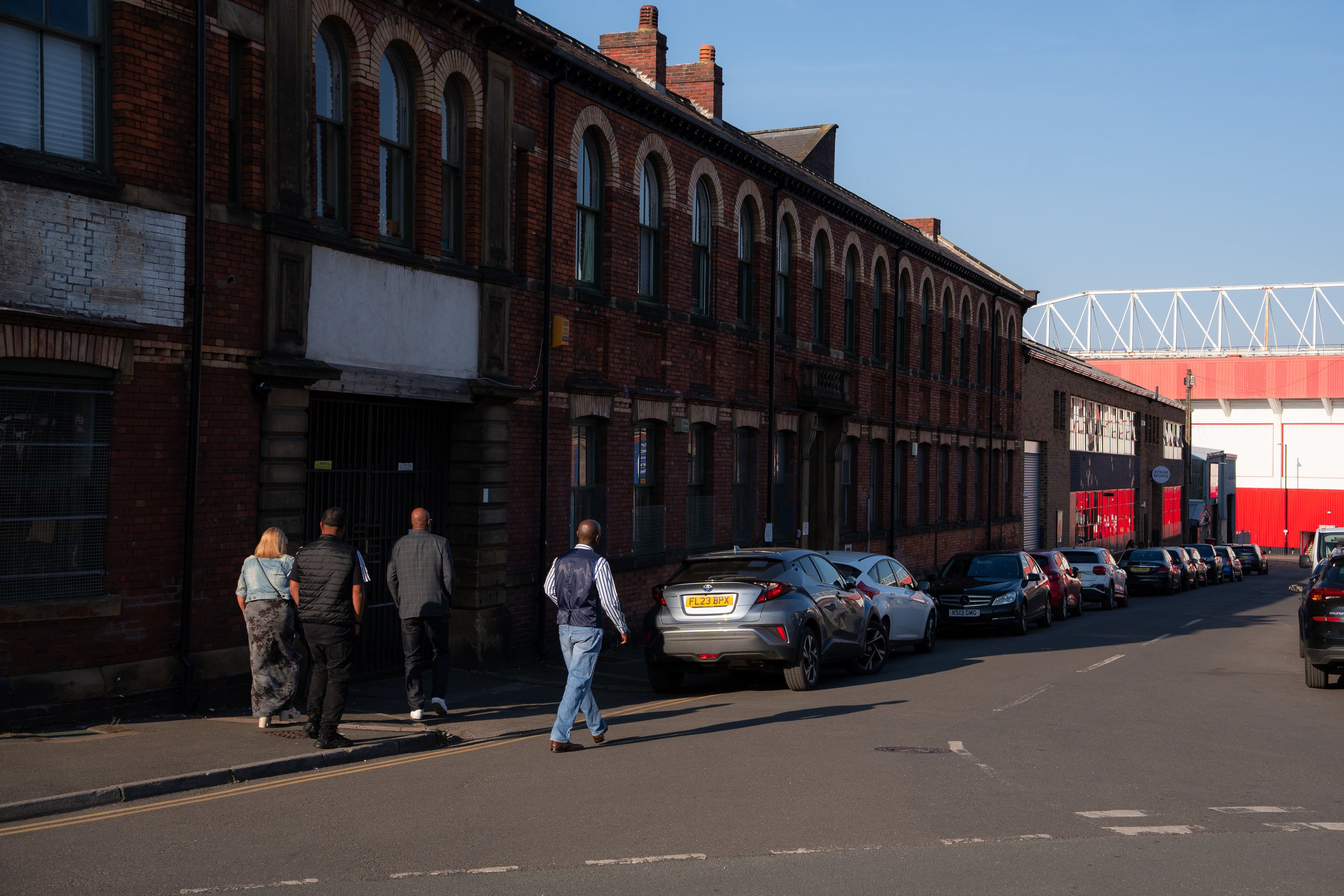 Group of five people walking along a city street next to parked cars and old brick buildings under a clear blue sky.