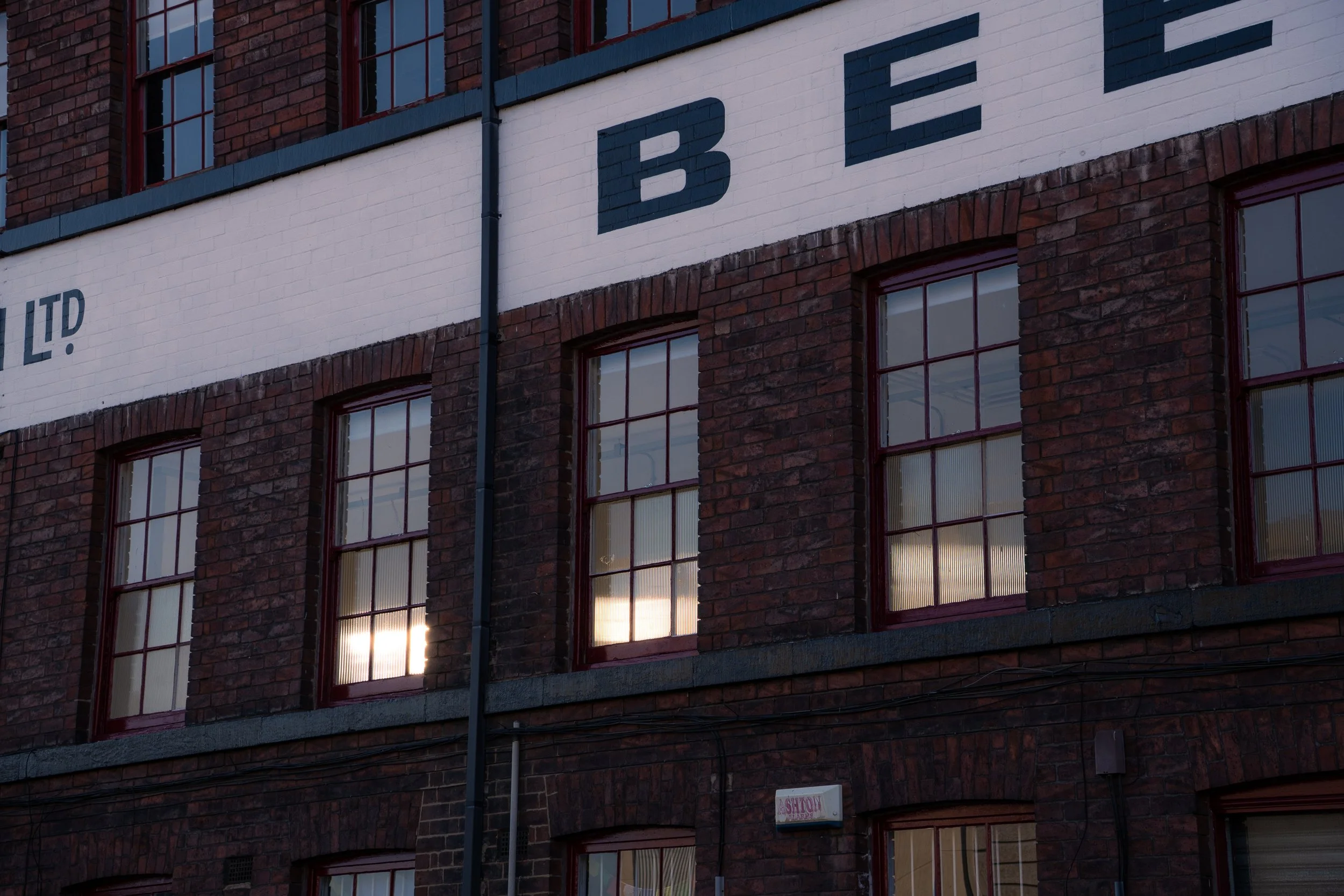 Close-up of a brick building with multiple windows. The building has a large white sign with partial dark blue letters that spell 'BE'. The windows reflect the sky and sunset. A small sign at the bottom reads 'ASHUT'.