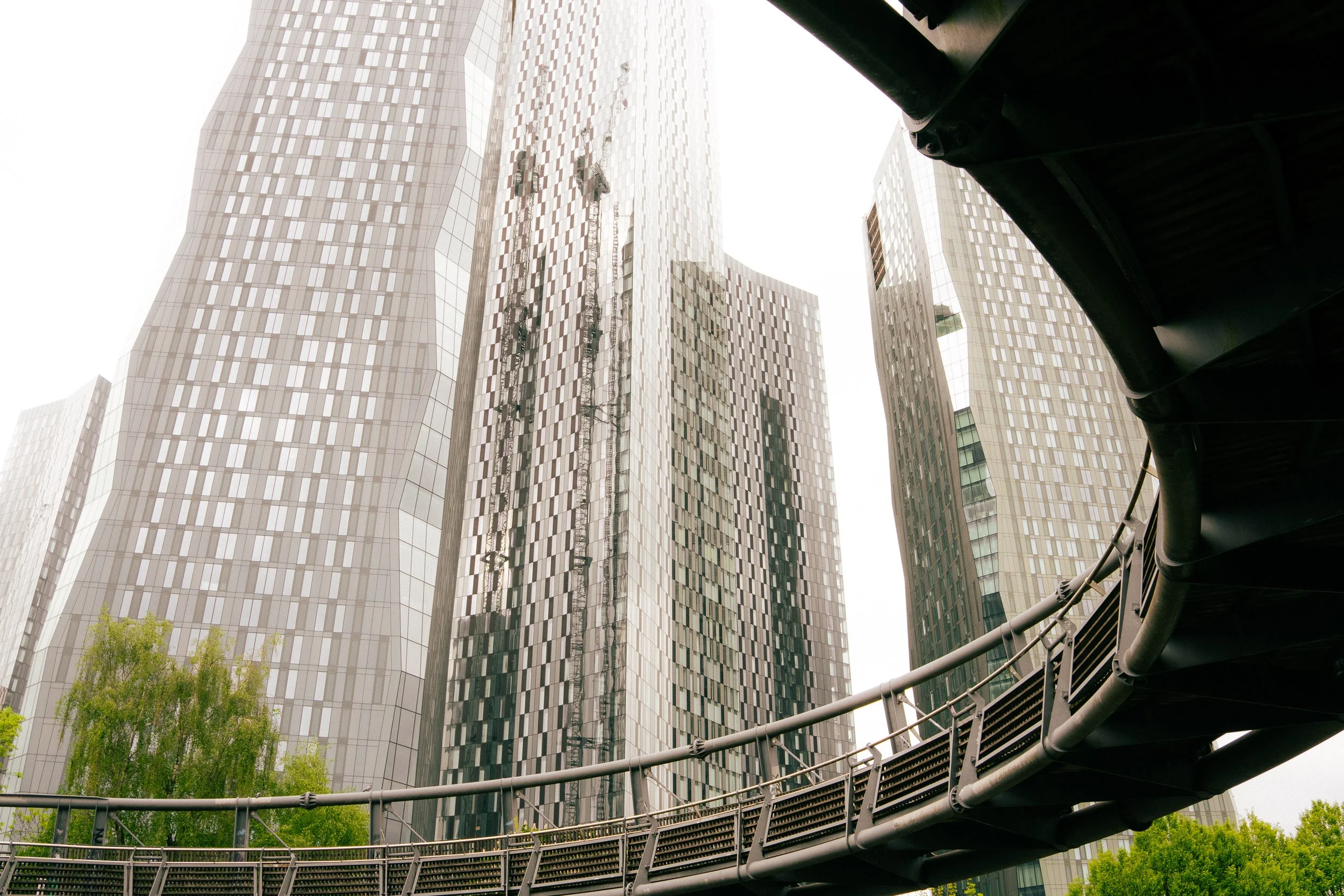 Low angle view of modern glass skyscraper buildings with curved and reflective surfaces, partly obscured by a dark overhang and metallic railing in the foreground.