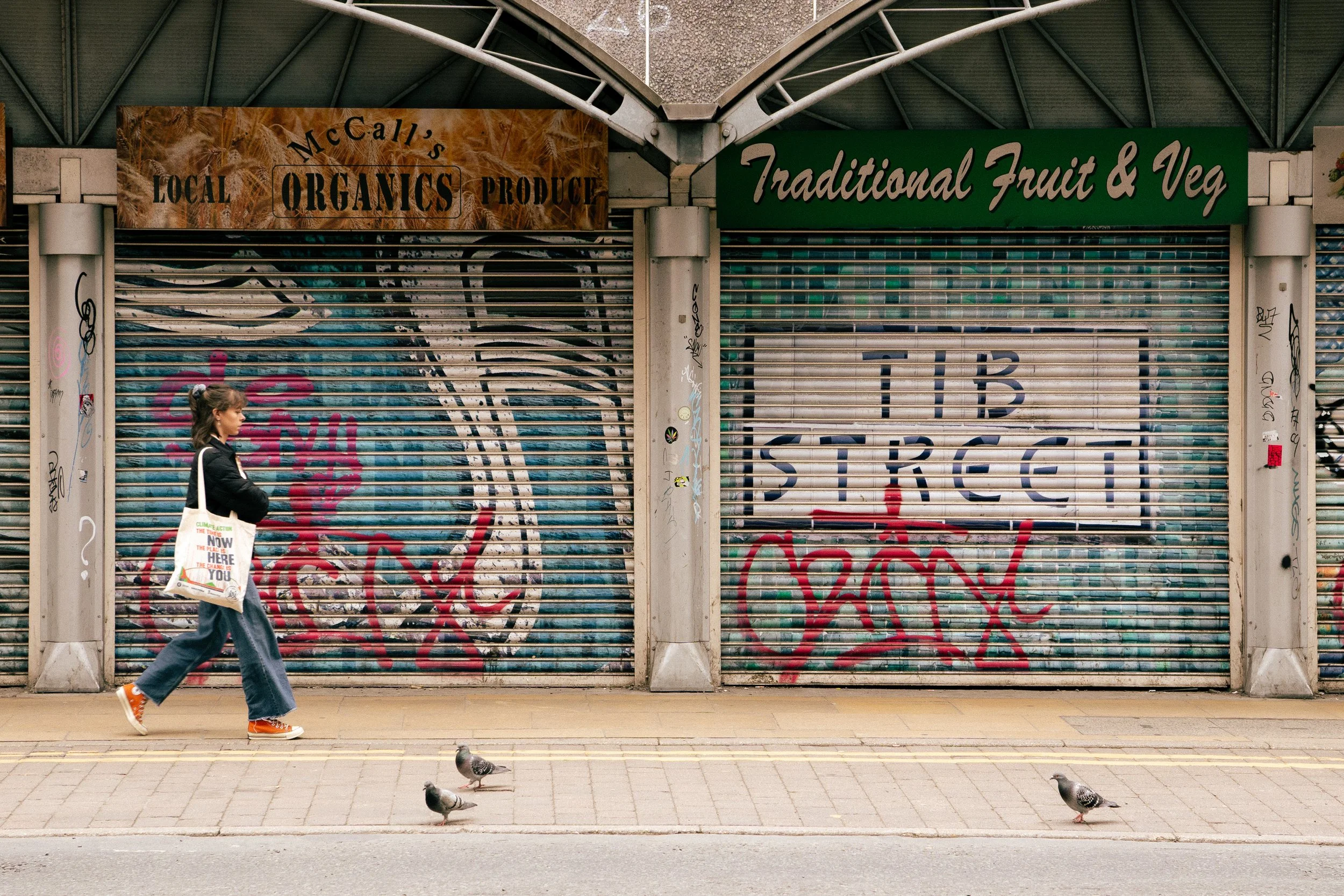 Closed storefront with graffiti on metal shutters, two signs above: one advertising local organic produce and another for traditional fruit and vegetables; a woman walking past with grocery bag, pigeons on the sidewalk.