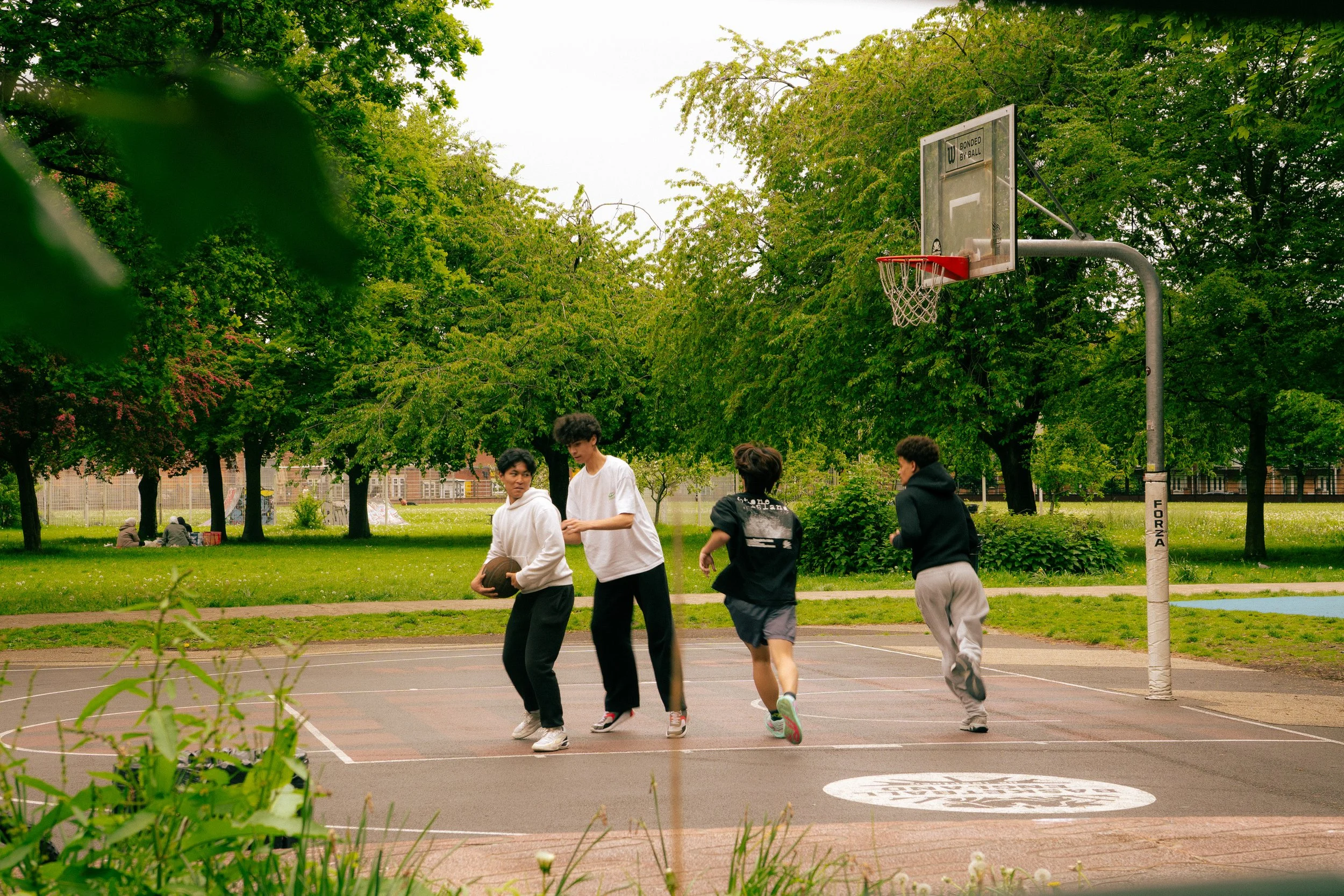 Group of young men playing basketball on an outdoor court surrounded by green trees with a park in the background.