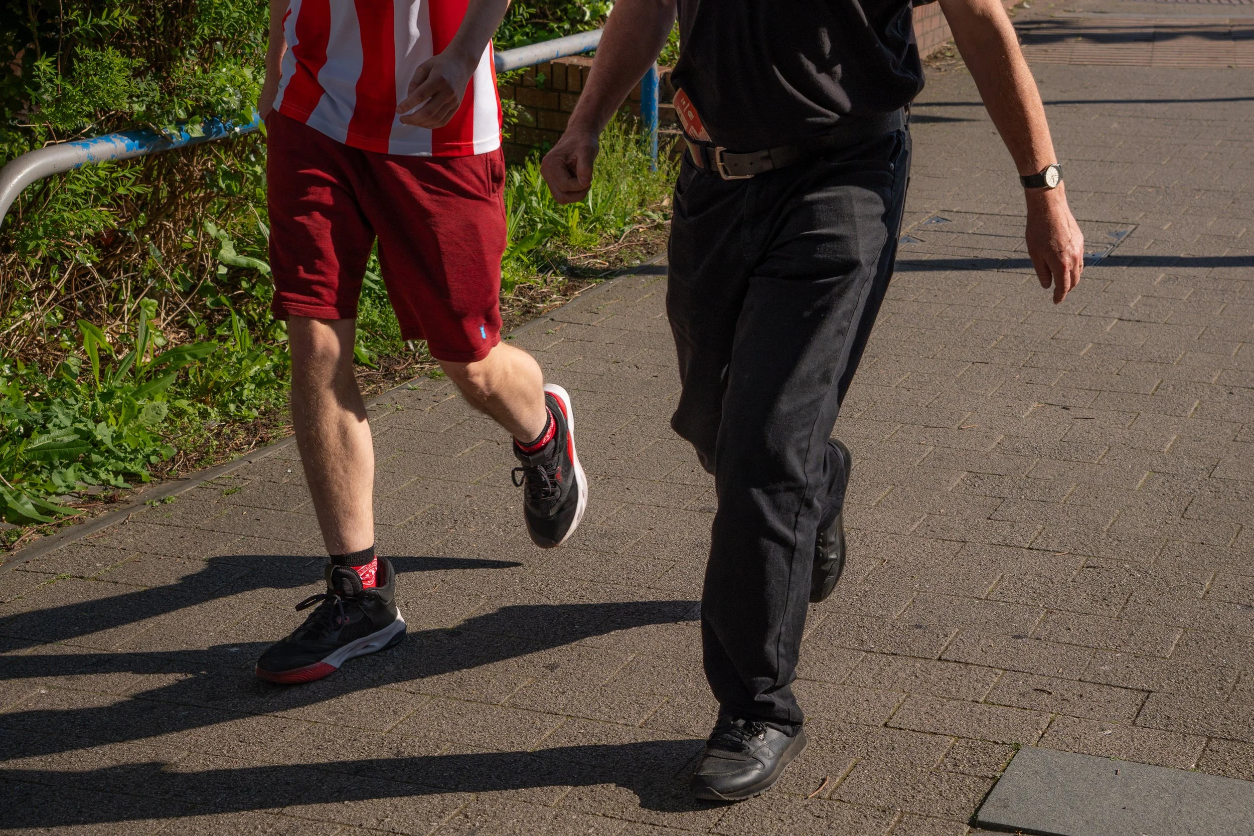 A person in red shorts and a striped shirt jogging or walking on a paved sidewalk, with another person in black pants and shirt walking nearby. Green bushes are visible on the left side of the image.