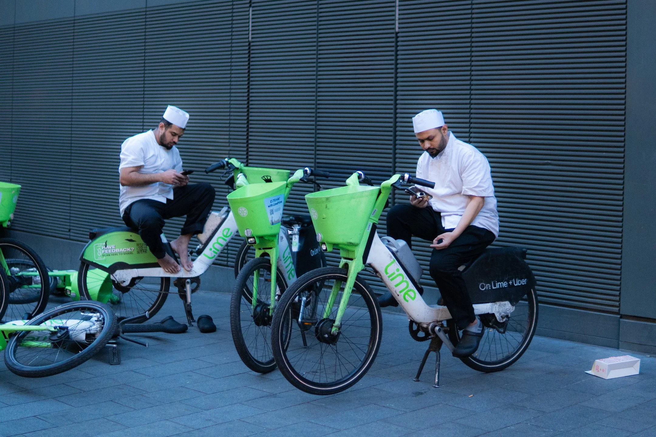Two men in white uniforms and hats sitting on Lime electric bikes, distracted by their phones, against a modern dark gray wall, with other bikes and a box on the ground nearby.