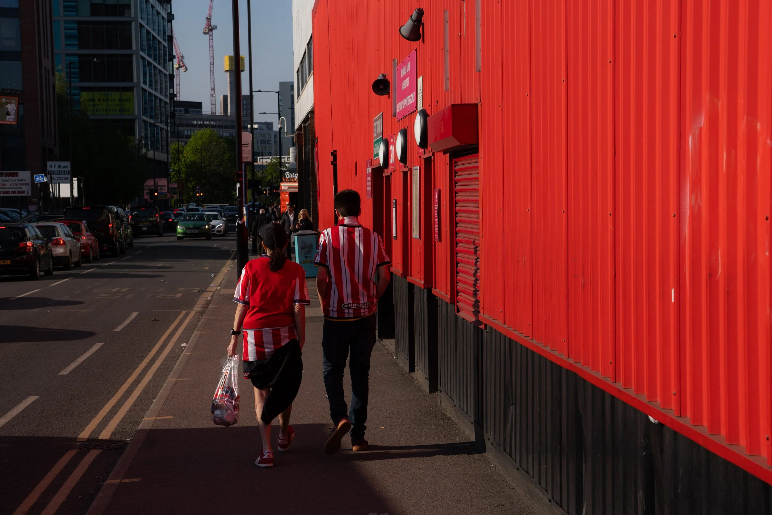 Two people walking past a bright red building in an urban street, with cars parked along the street and modern glass buildings in the background.