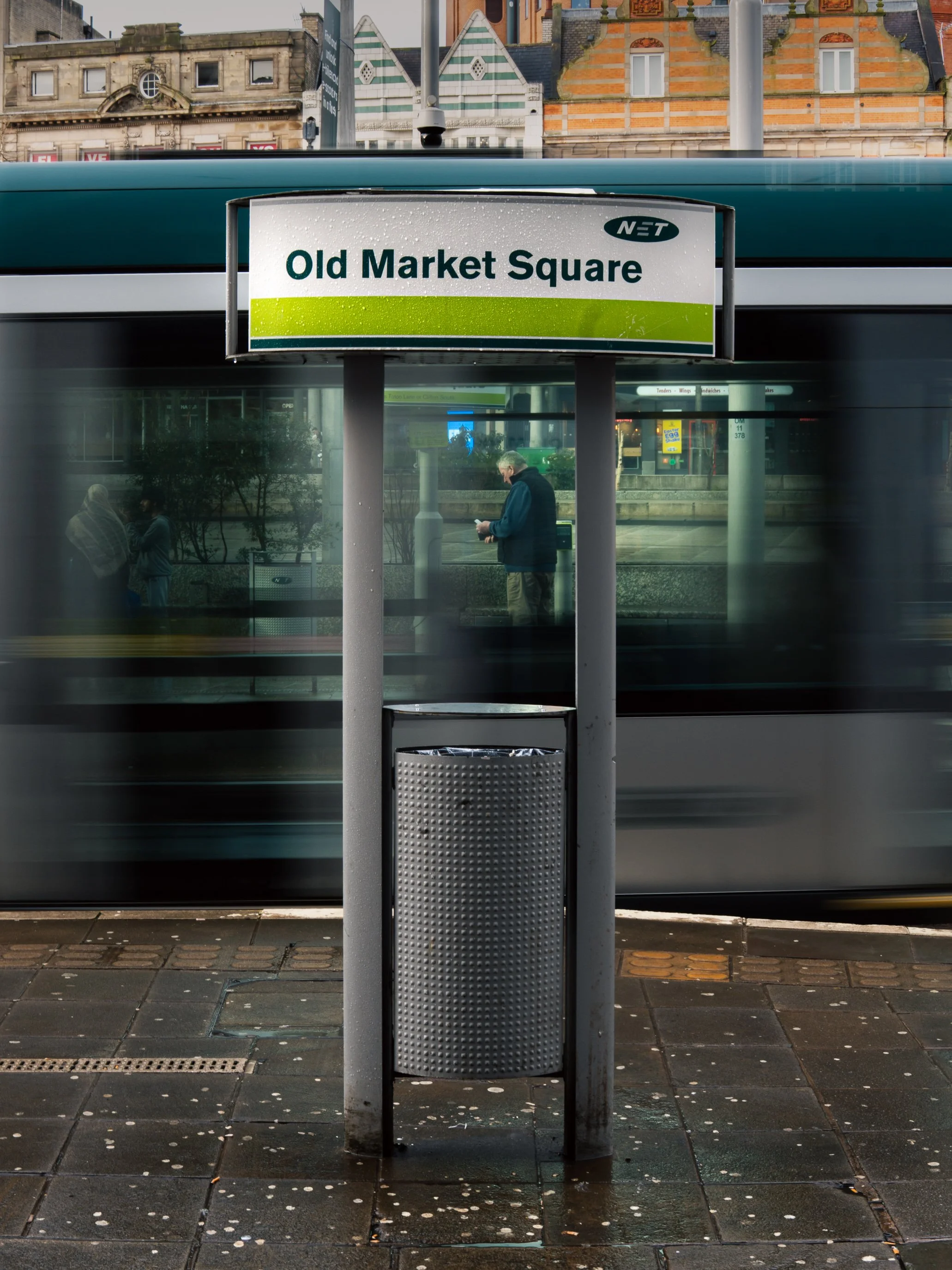 A city street scene featuring a bus stop sign for Old Market Square with a person standing beneath it, reflections of buildings and people visible in the background, and a moving bus passing by in front of the bus stop.