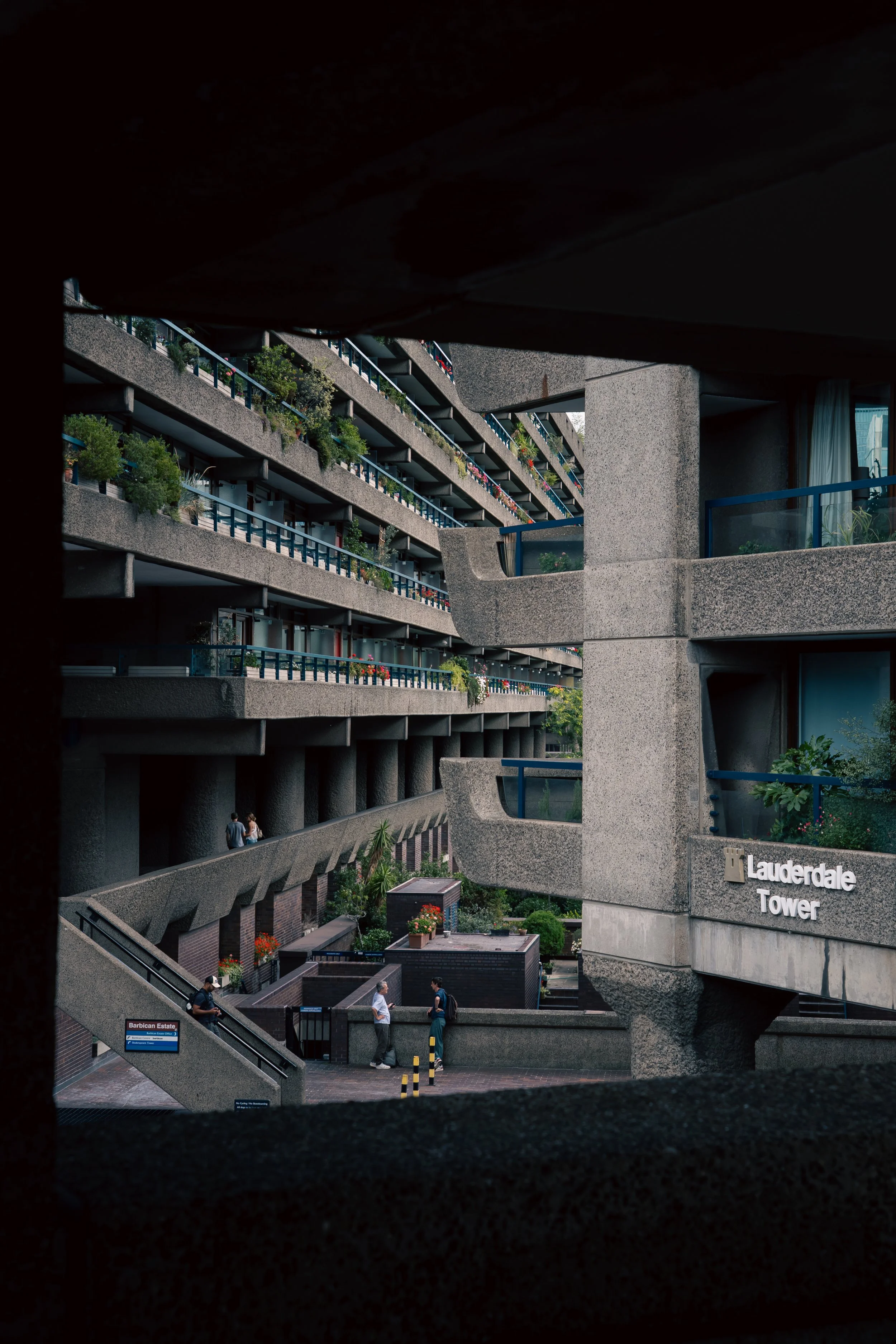 Multi-story concrete apartment buildings with balconies and plants, viewed through a dark frame, with a sign that reads 'Lauderdale Tower' and people walking on the sidewalk below.