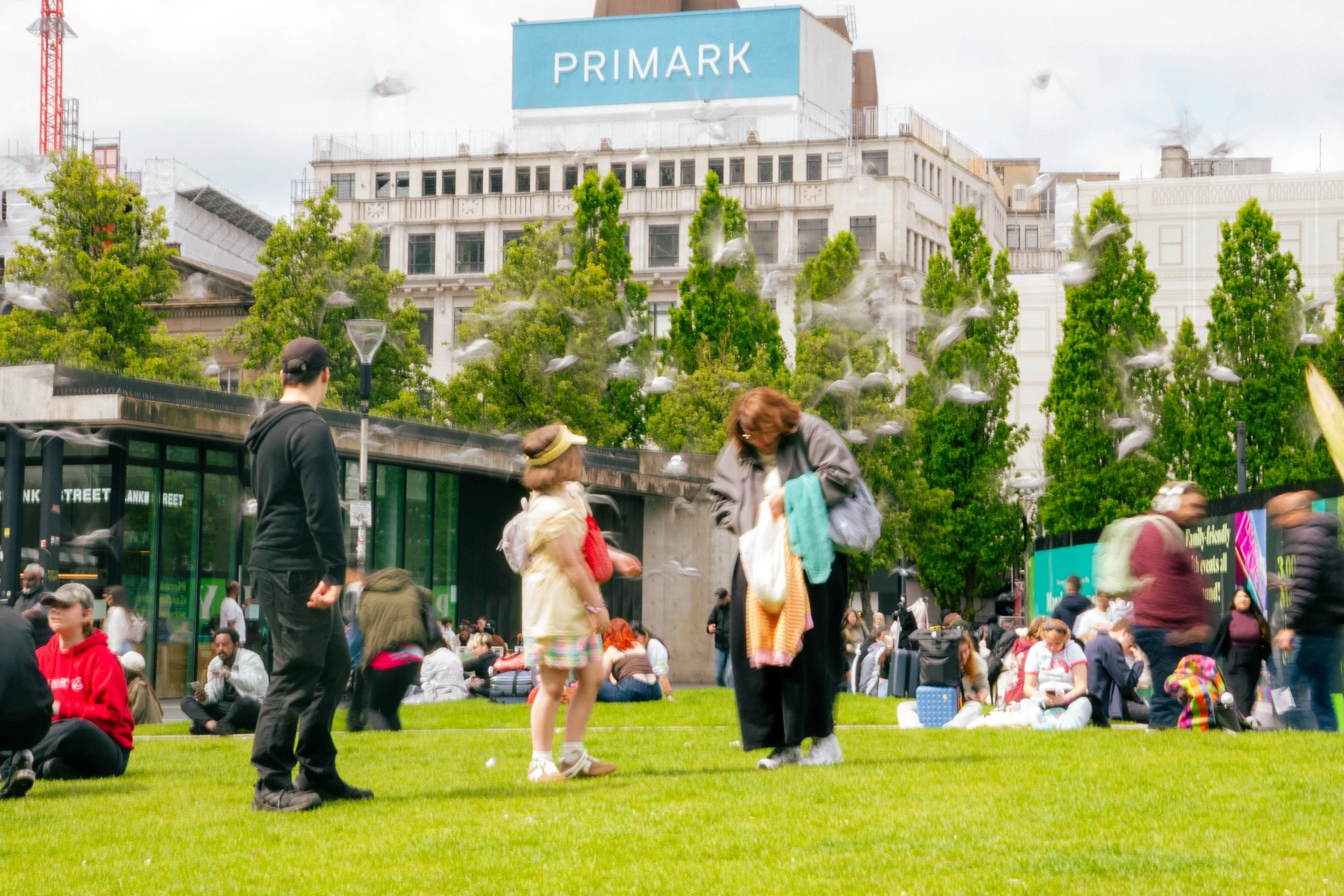 People gathered on a grassy area in an urban setting, with some sitting and others standing, amidst a crowd. In the background, there are tall trees, a city building with a 'Primark' sign on top, and a modern glass structure. The scene appears lively