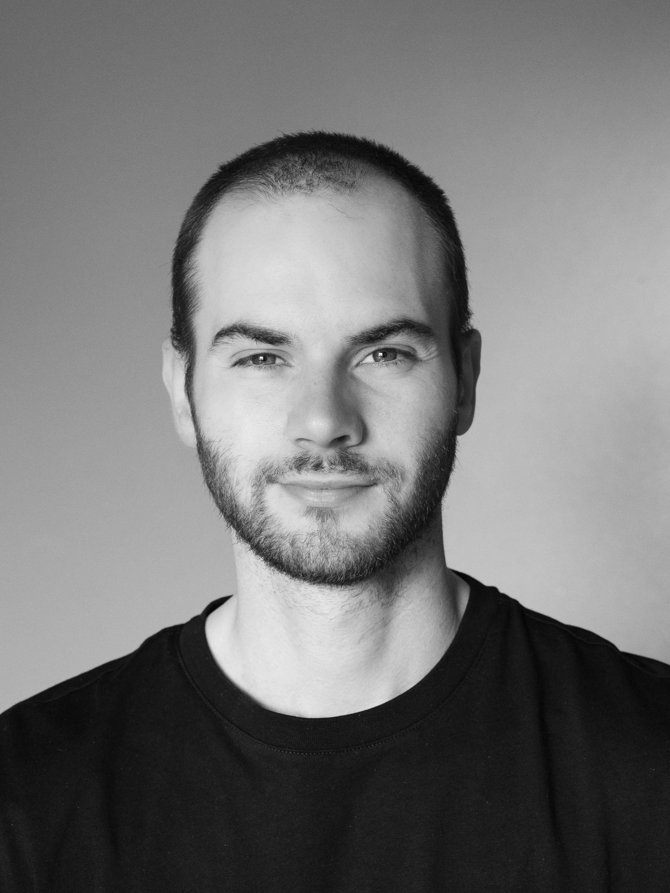 Black and white portrait of a smiling young man with short hair and facial hair, wearing a black shirt.