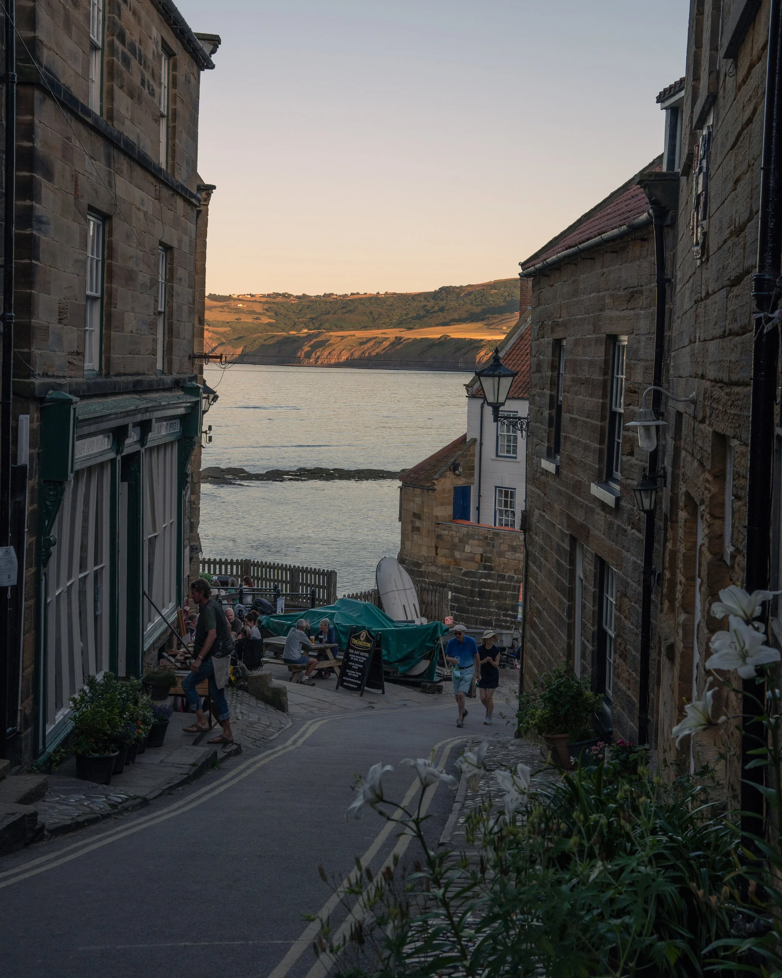 A street scene in a coastal town during sunset, with people sitting at outdoor tables, old stone buildings on either side, and a view of water and hills in the background.