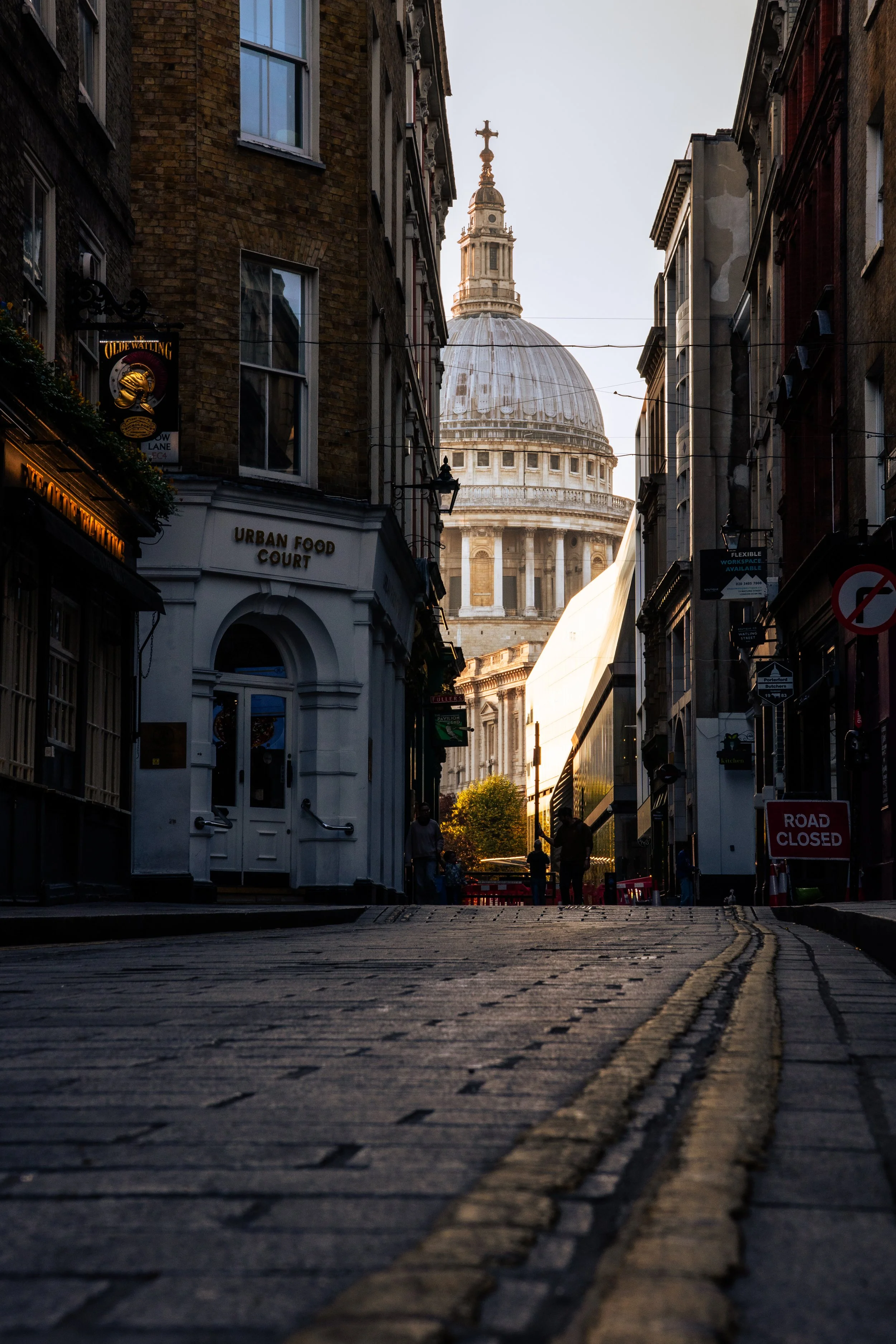 A street view in London with a view of St. Paul's Cathedral in the background during sunset. The street is lined with buildings with shops, including one with a sign reading 'Urban Food Court.' The street is closed to traffic, with a sign indicating 