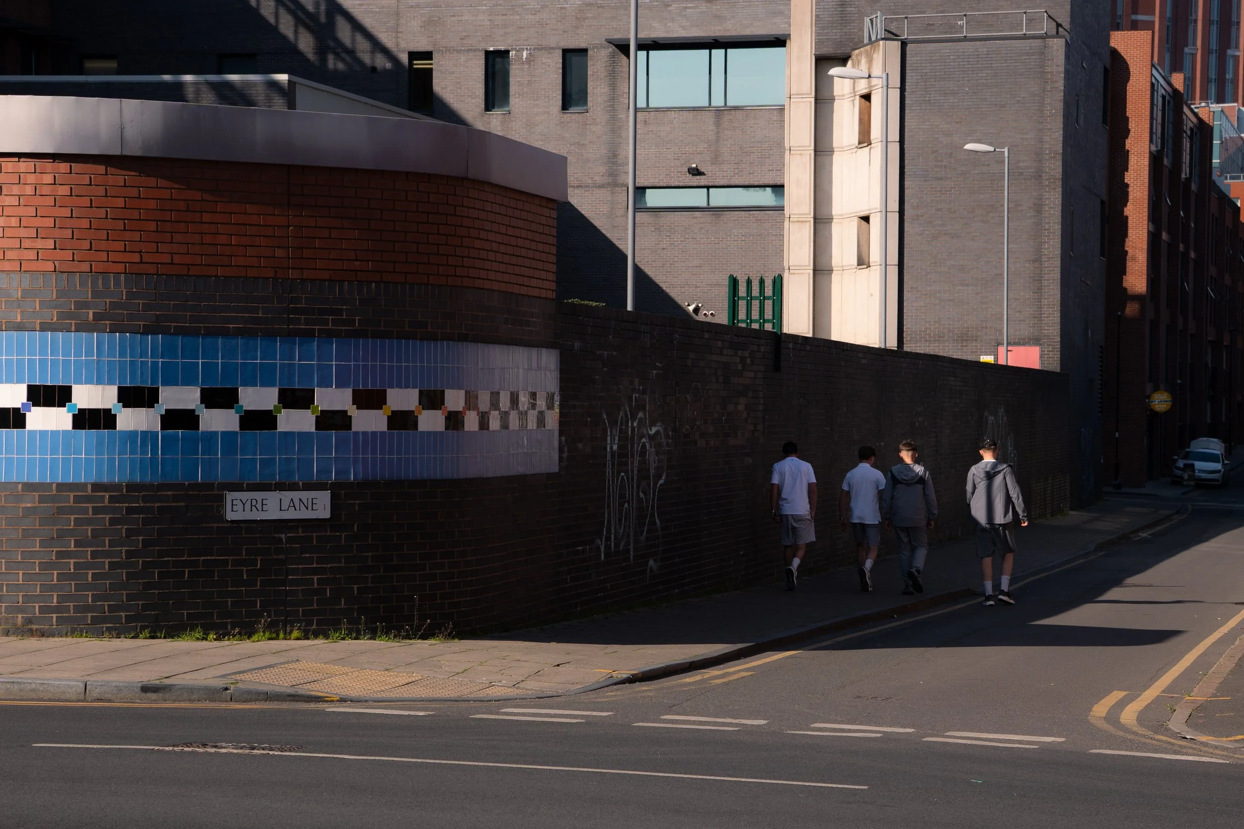 Four people walking along a sidewalk in front of a brick wall and modern buildings during daytime.