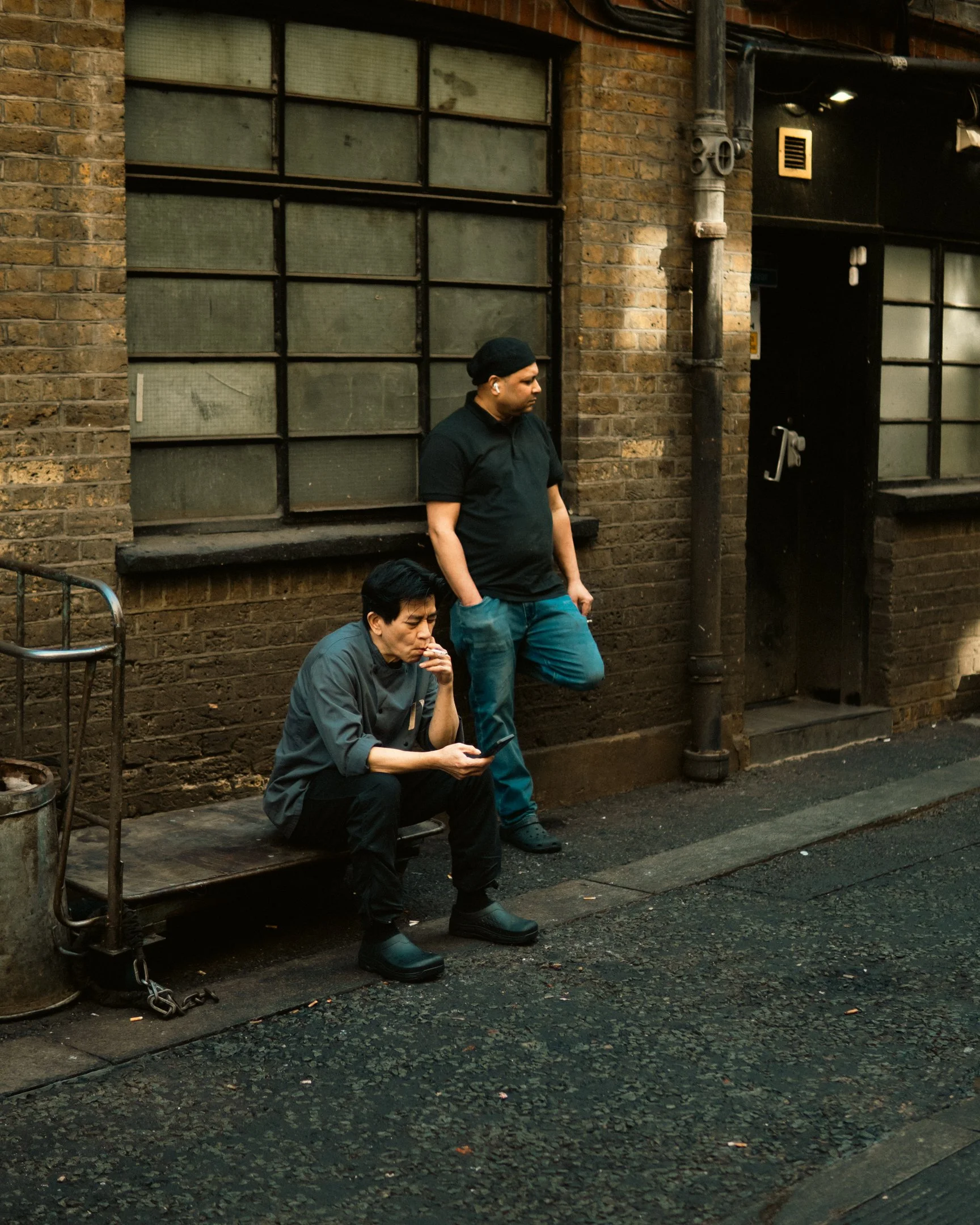 A man in black standing with hands in pockets and two seated men, one eating, outside a brick building with large windows and a black door.