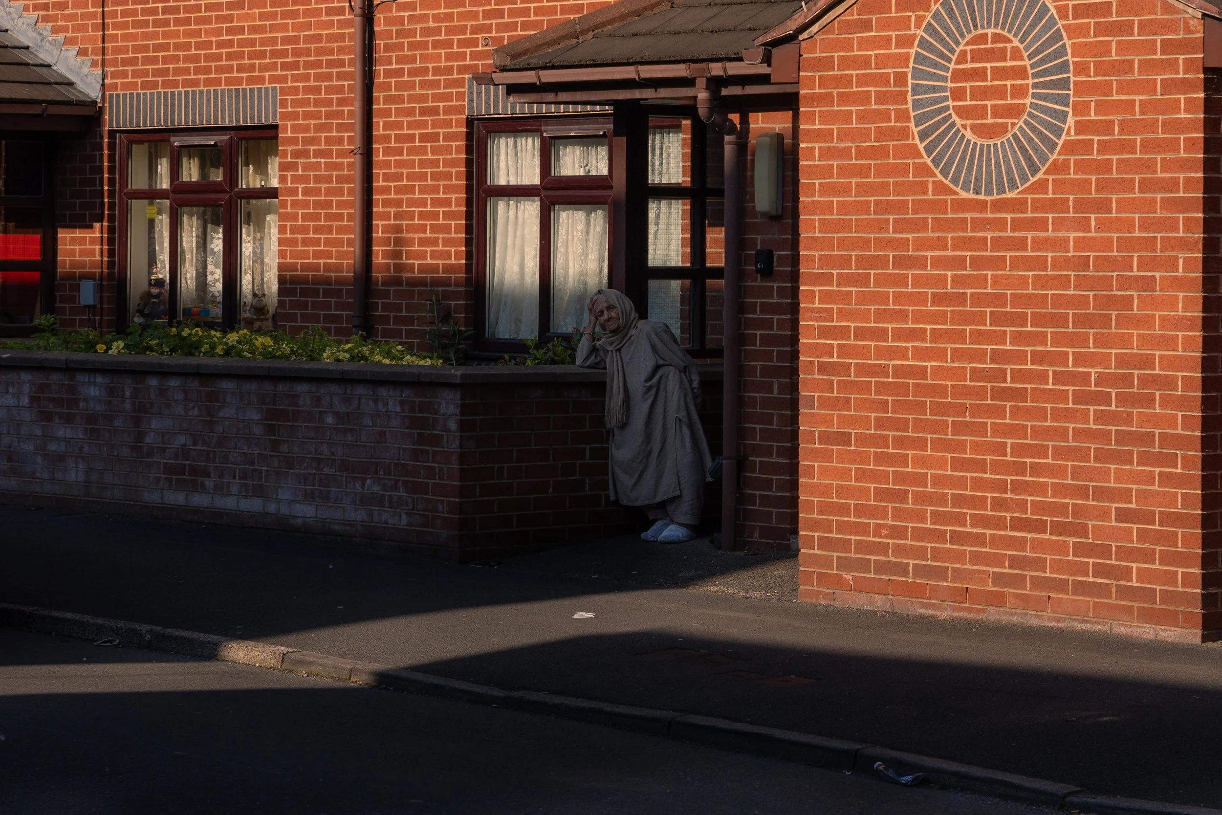A woman dressed in a long beige coat, gray headscarf, and slippers standing outside a red brick house, leaning against a brick wall, with a garden bed and windows behind her.