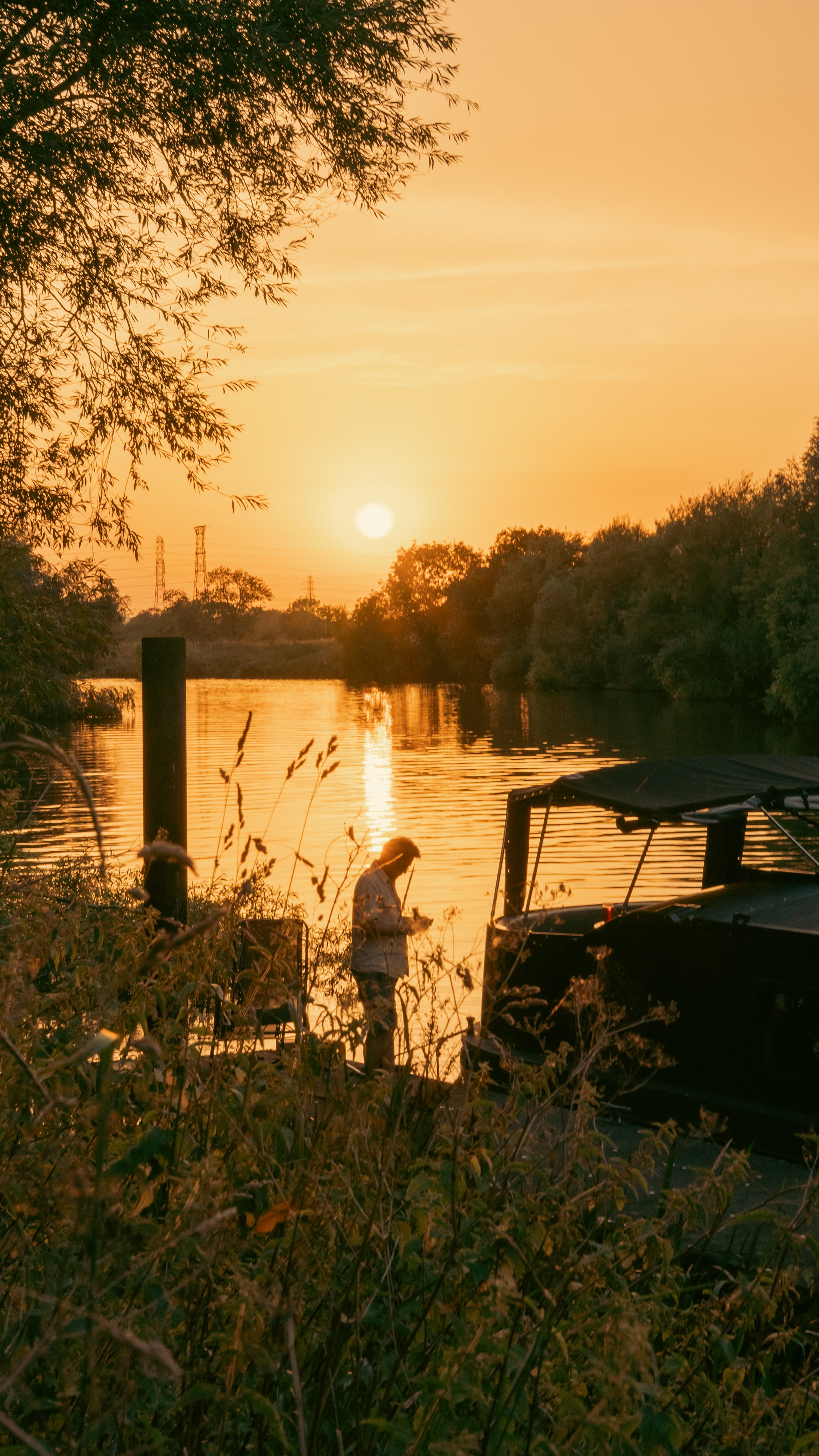 A person standing near a dock by a river during sunset, with trees and power lines in the background, and a boat in the foreground.