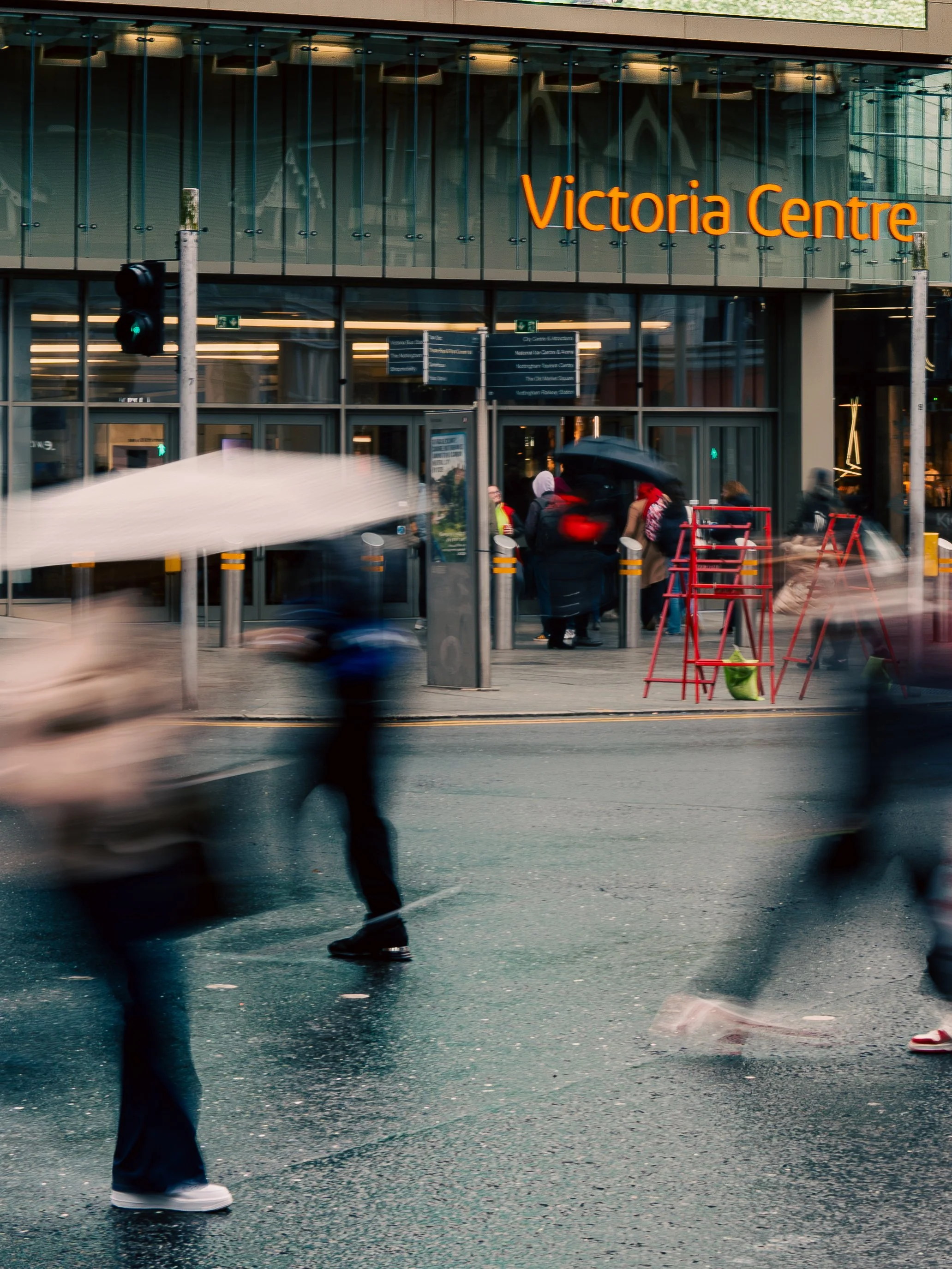 The exterior of Victoria Centre shopping mall with people walking by, some holding umbrellas on a rainy day, and motion-blurred pedestrians passing the camera.