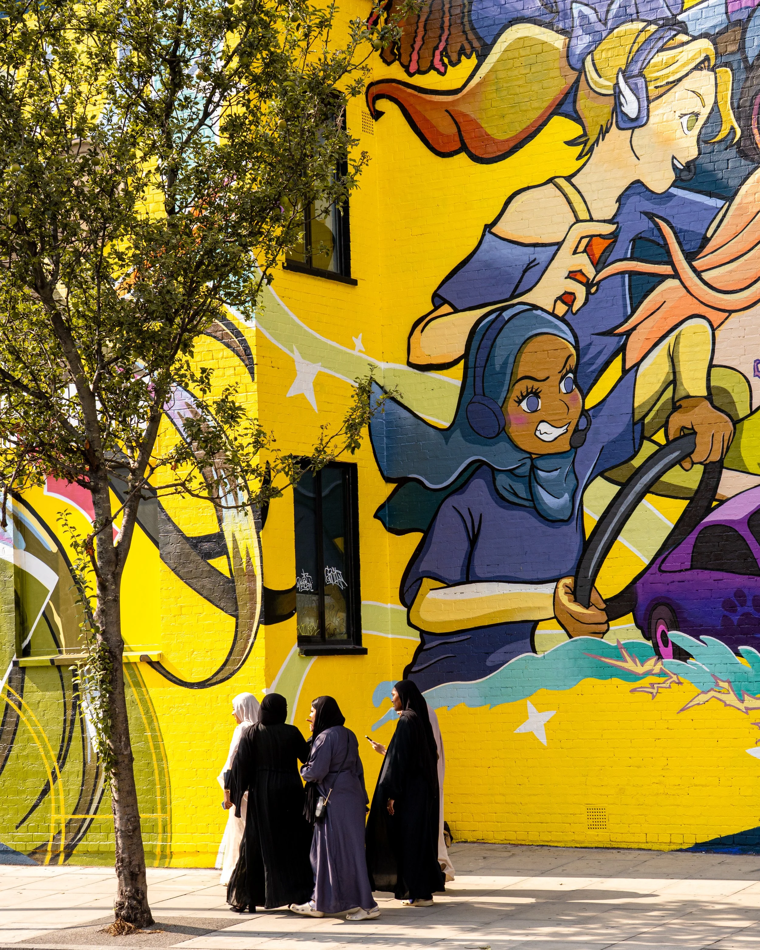 People in full-body black traditional robes walking past a large, colorful mural on a yellow brick wall, with a tree partially obscuring the artwork.