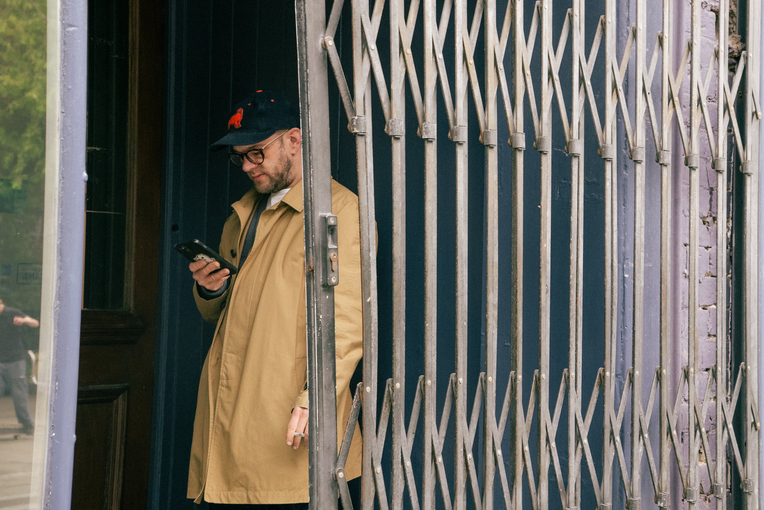 A man wearing glasses, a tan coat, and a black cap with red embroidered design standing behind a metal gate, looking at his phone.