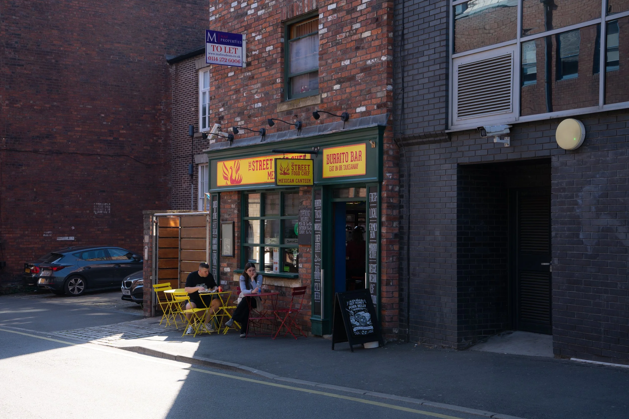 A small Mexican restaurant named The Street Me, with outdoor yellow and red chairs, where two people are sitting and eating outside. The restaurant has yellow signs with red text and a green door, situated on a brick building with nearby parked cars.