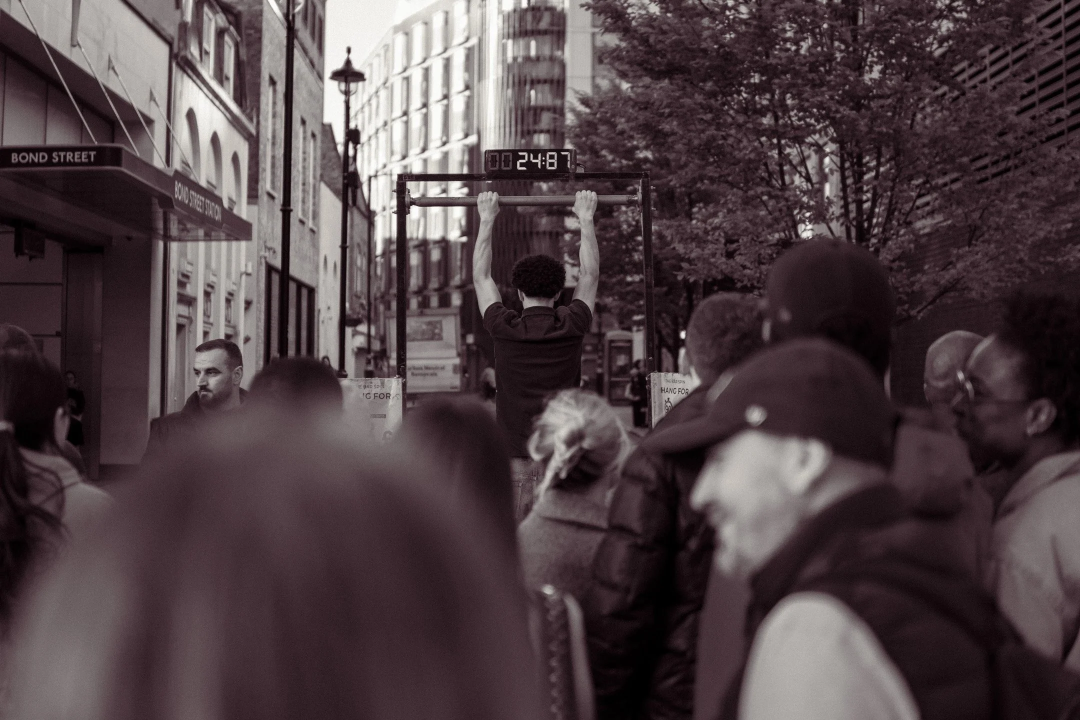 A person with curly hair is doing a pull-up on a bar in a street setting. A digital timer is above the bar showing 24:87. There are several people in the foreground and buildings lining the street, with a street sign named 'Bond Street' visible on th