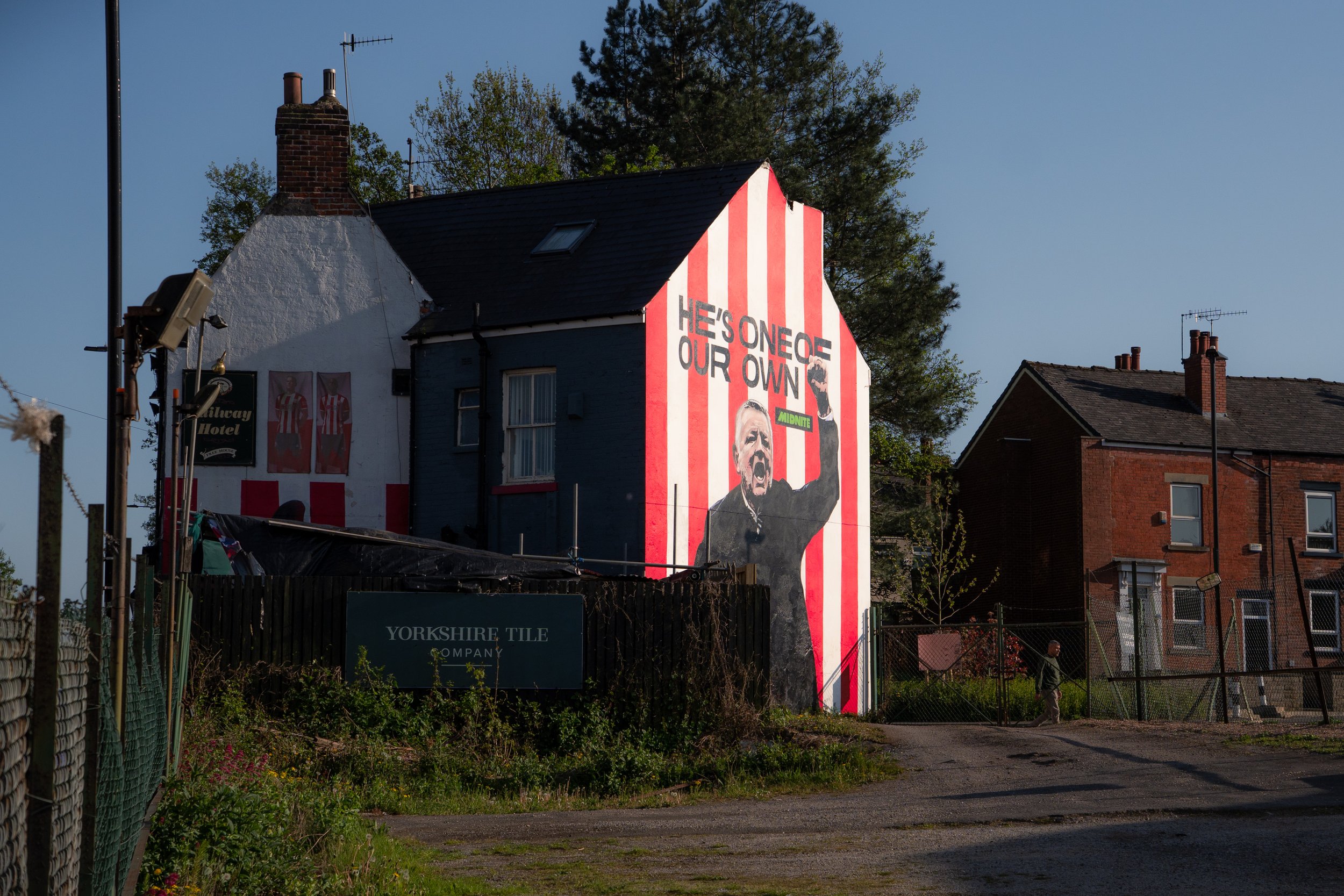 Large painted mural on side of building with red and white vertical stripes, depicting a man with his fist raised and saying, 'HE'S ONE OF OUR OWN,' with the word 'MIDNITE' in green.
