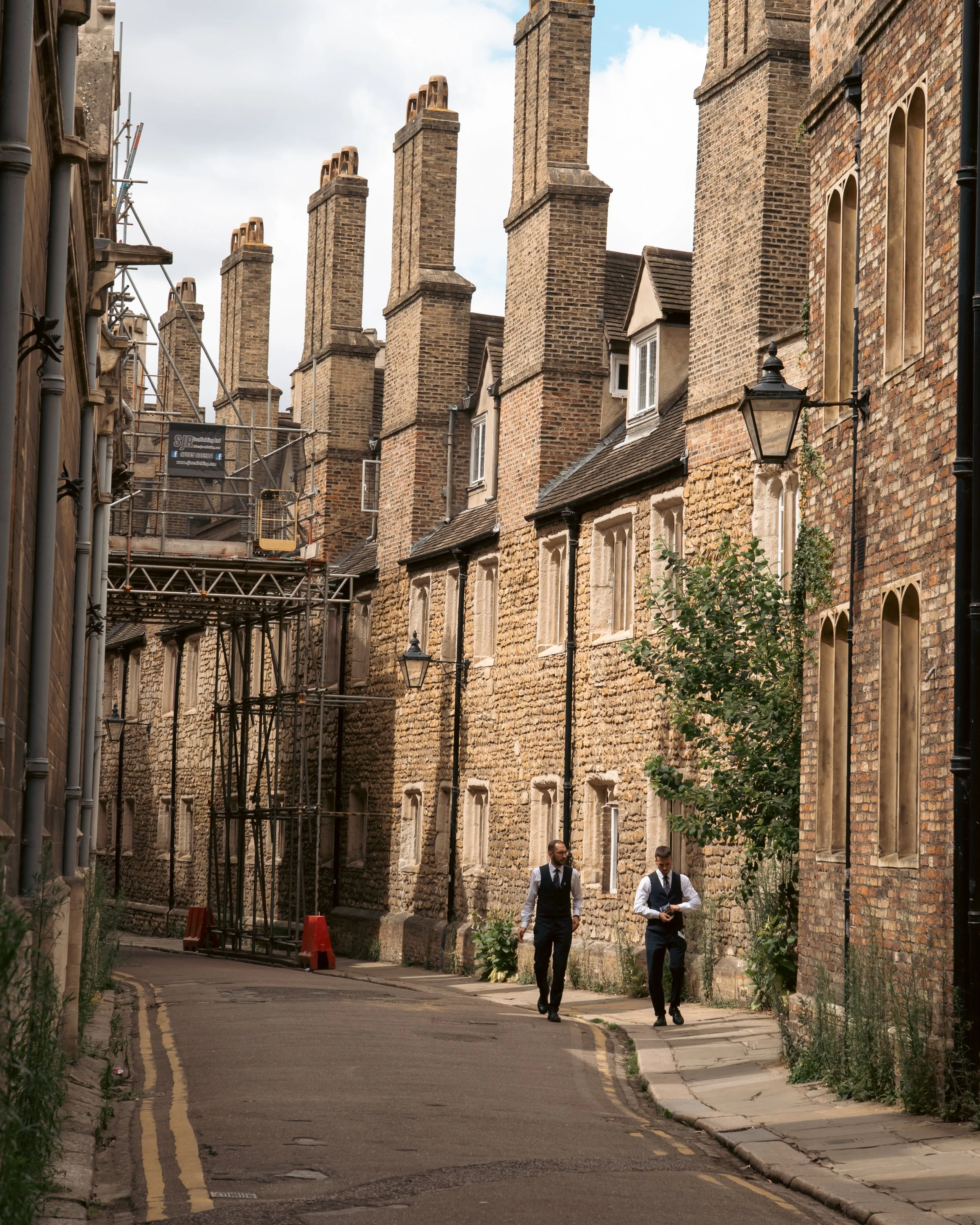 Two men dressed in formal business attire walking down a narrow, brick-lined street. The street is lined with old brick buildings with tall chimneys. There is a street lamp on the right side and construction scaffolding on the left side. One man is l