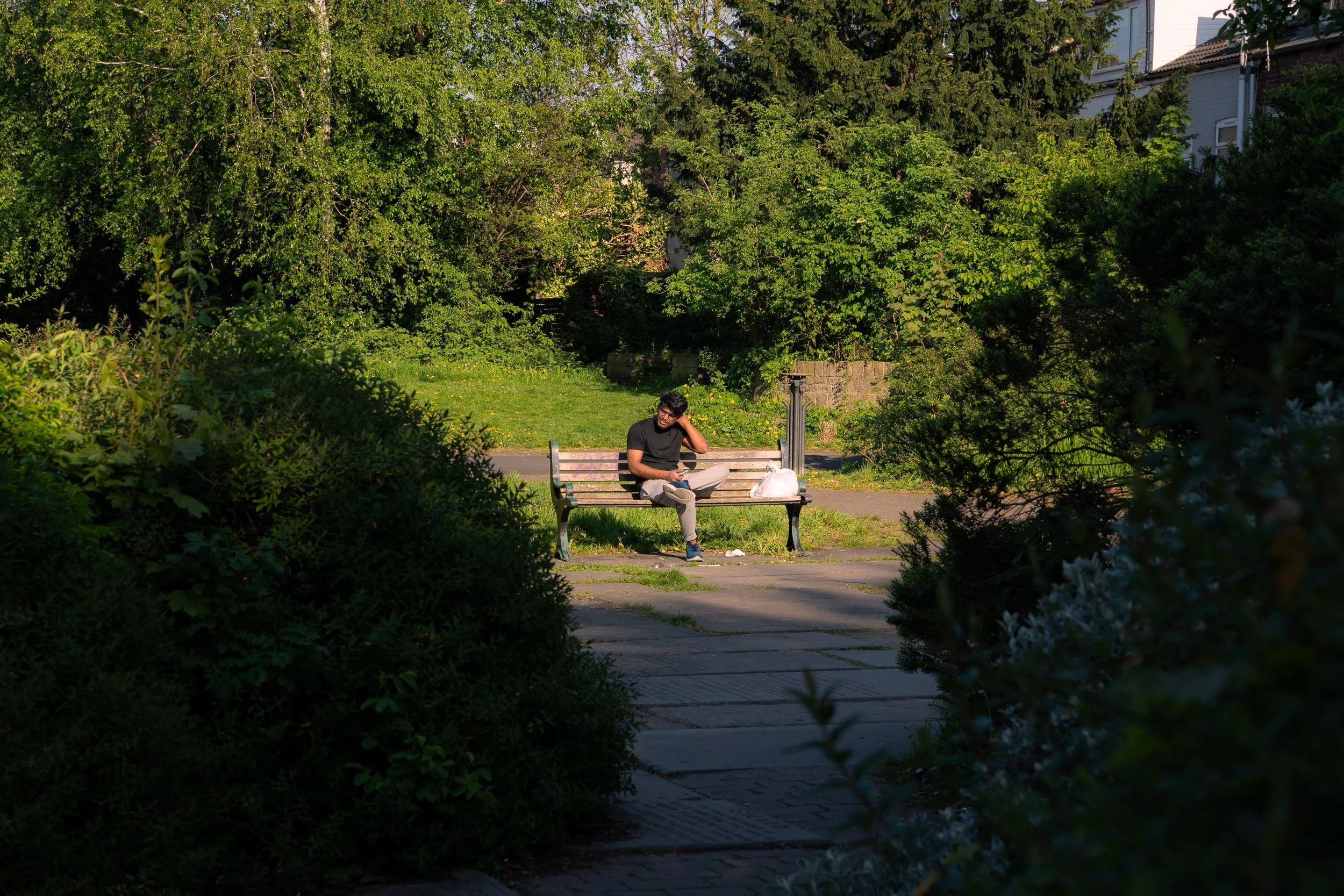 A man sitting on a park bench in a green, leafy park with trees and bushes surrounding him, holding a phone, with a bag on the bench beside him.