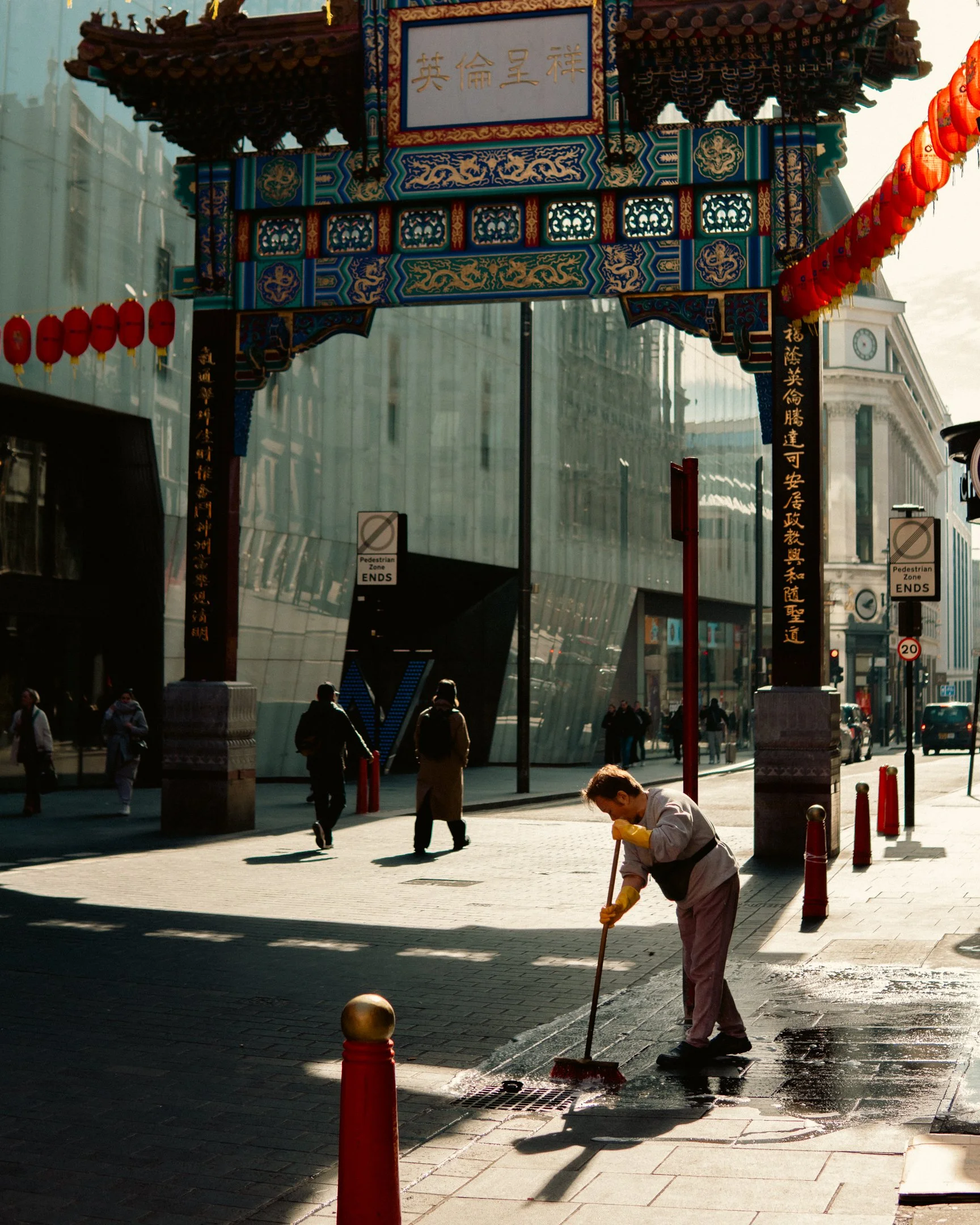 A man cleaning a sidewalk with a broom in an urban area, with a traditional Chinese gateway arch overhead.
