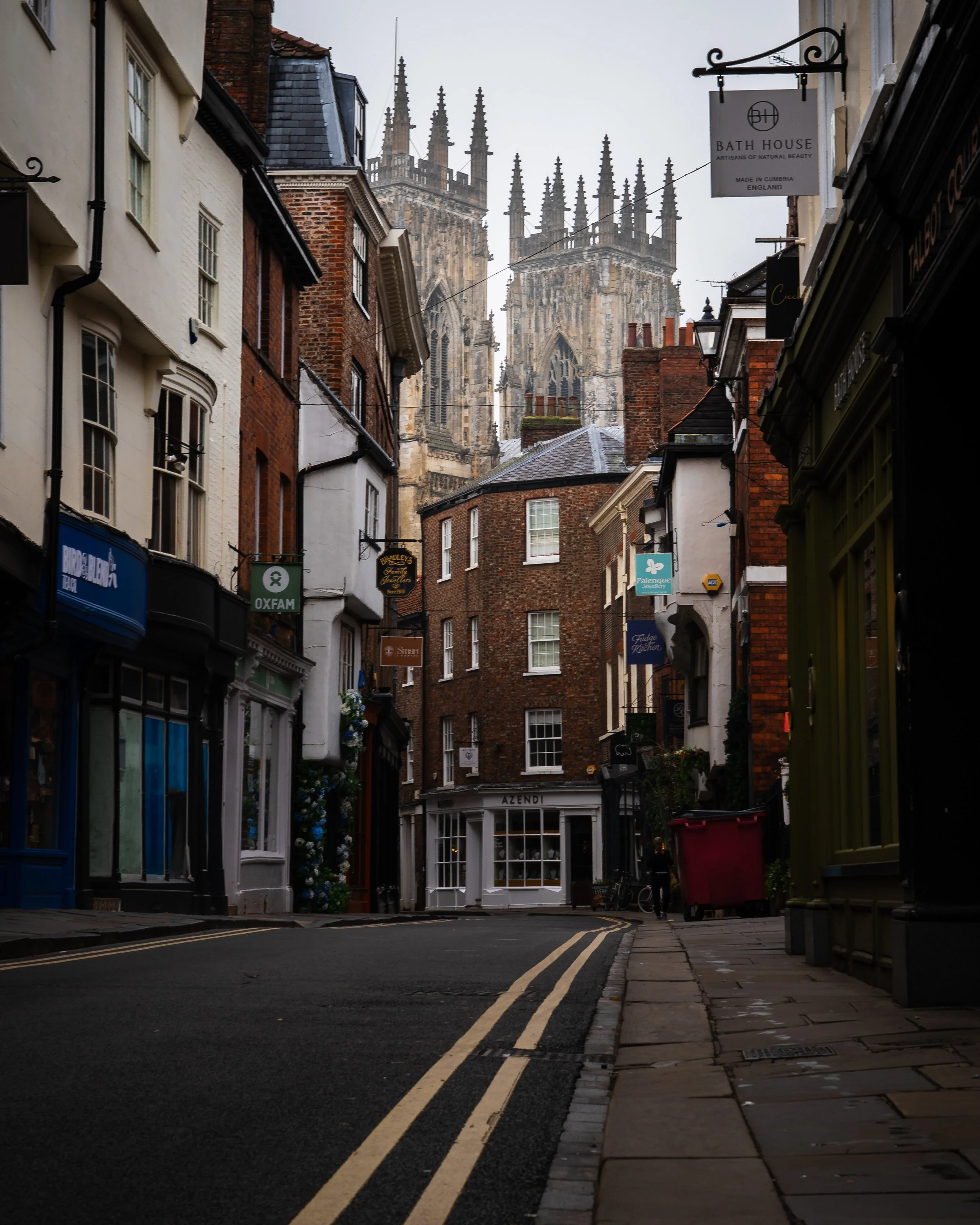 A narrow street in an old European town with shops on both sides and a large cathedral with tall spires in the background.
