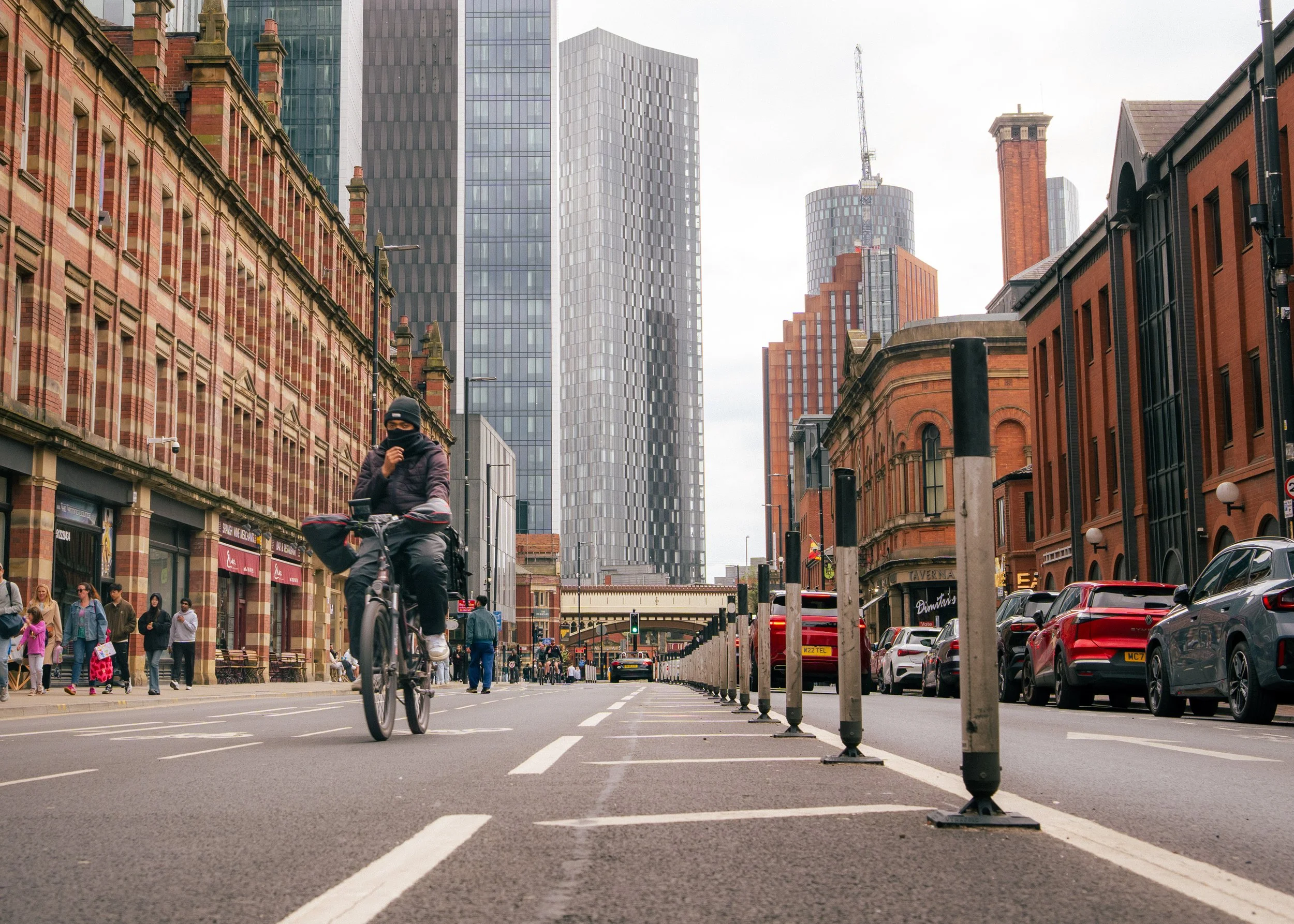 City street scene with pedestrians and a cyclist in the foreground, modern high-rise buildings, vintage brick buildings, parked cars, street lamps, and a cloudy sky.